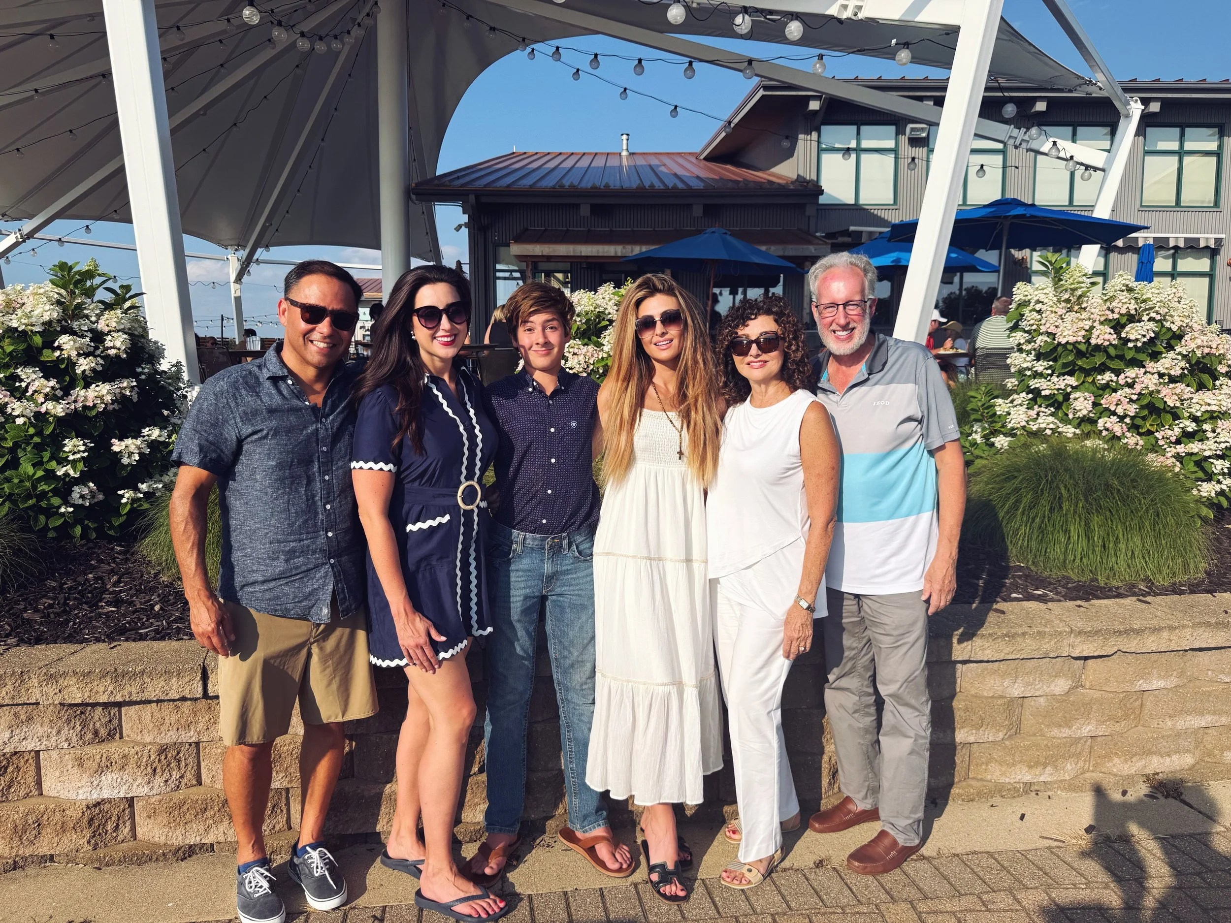 Group of six people standing outdoors on a sunny day, posing for a photo, with a building, umbrellas, string lights, and blooming bushes in the background.