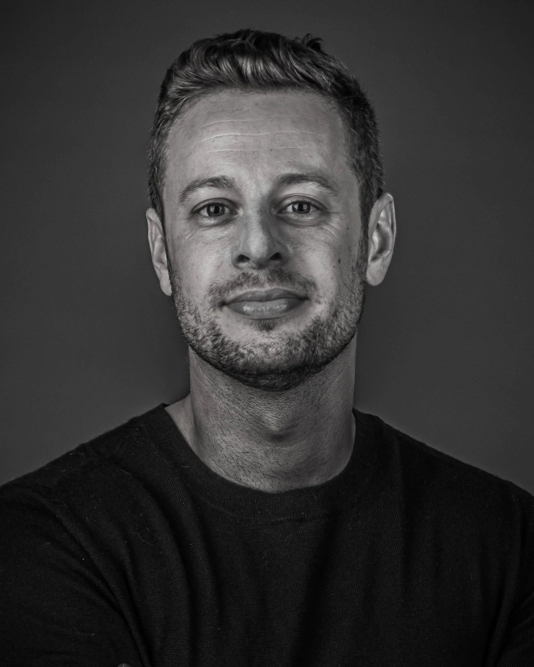 Close-up black and white portrait of a young man with short hair and a slight smile, wearing a dark sweater.