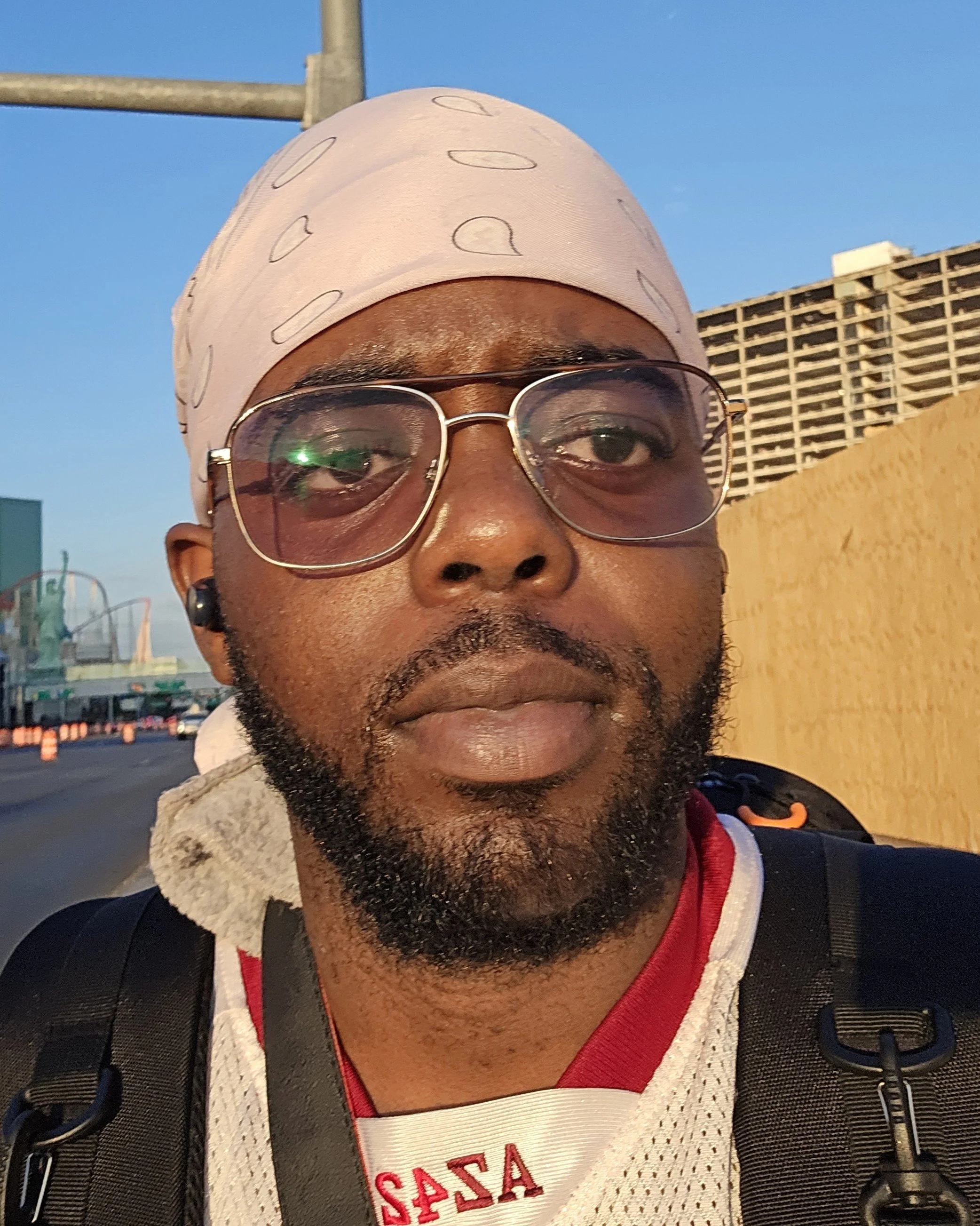 A man wearing glasses, a light-colored bandana, and a sports jersey takes a selfie outdoors, with a blue sky and some buildings in the background.