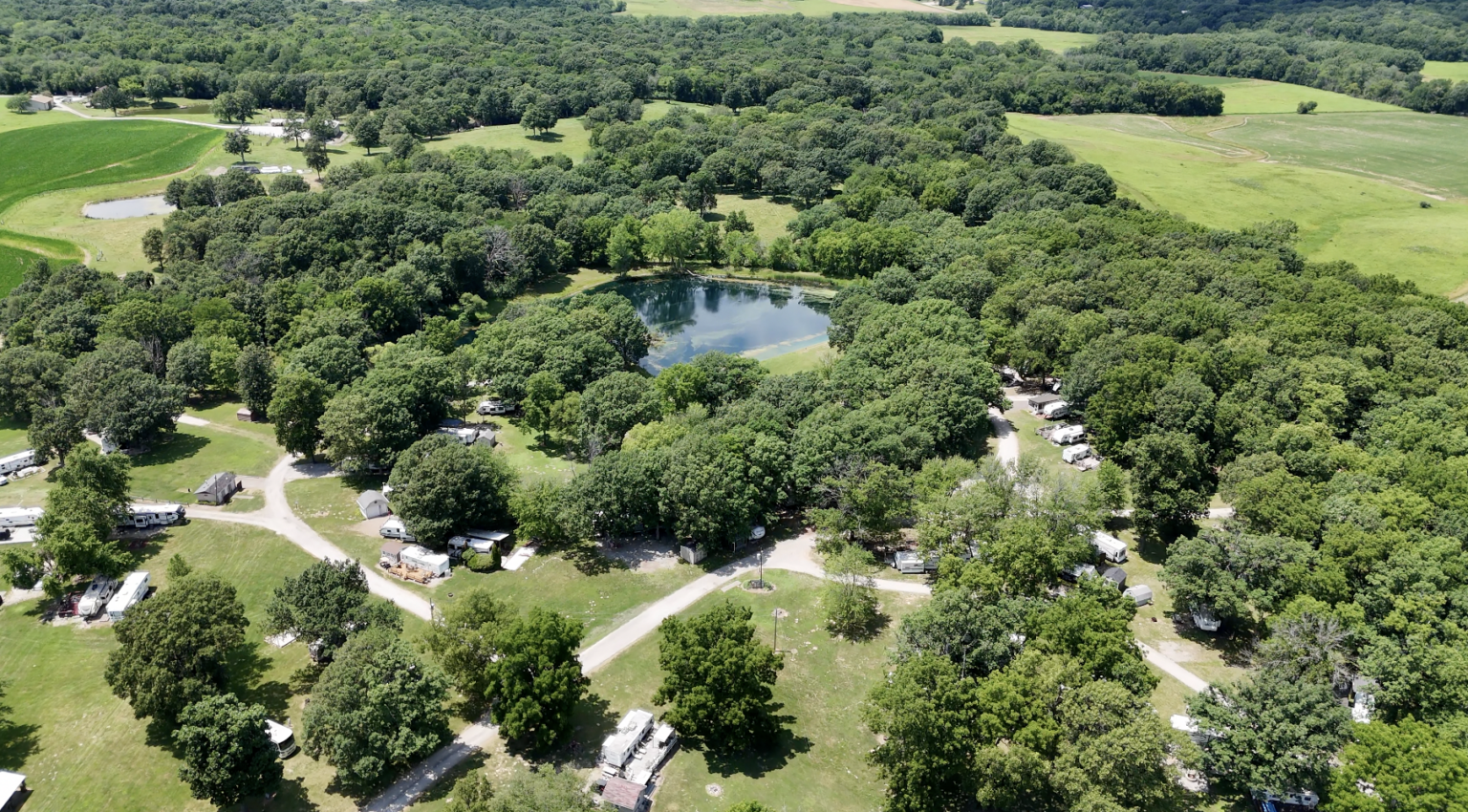 Aerial view of a wooded campground with camper trailers and caravans, small lakes, and surrounding green fields.