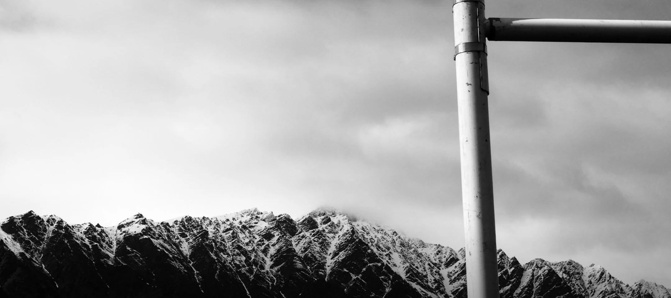 Black and white photo of rugged snow-capped mountains with a cloudy sky in the background. A metal pipe structure is visible on the right side of the image.