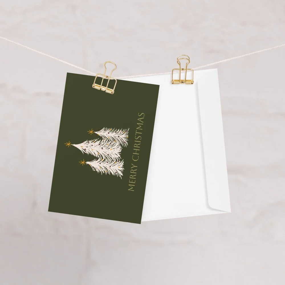A holiday greeting card hanging on a clothesline with gold paper clips, featuring a white Christmas tree illustration and the words "Merry Christmas" on a dark green background.