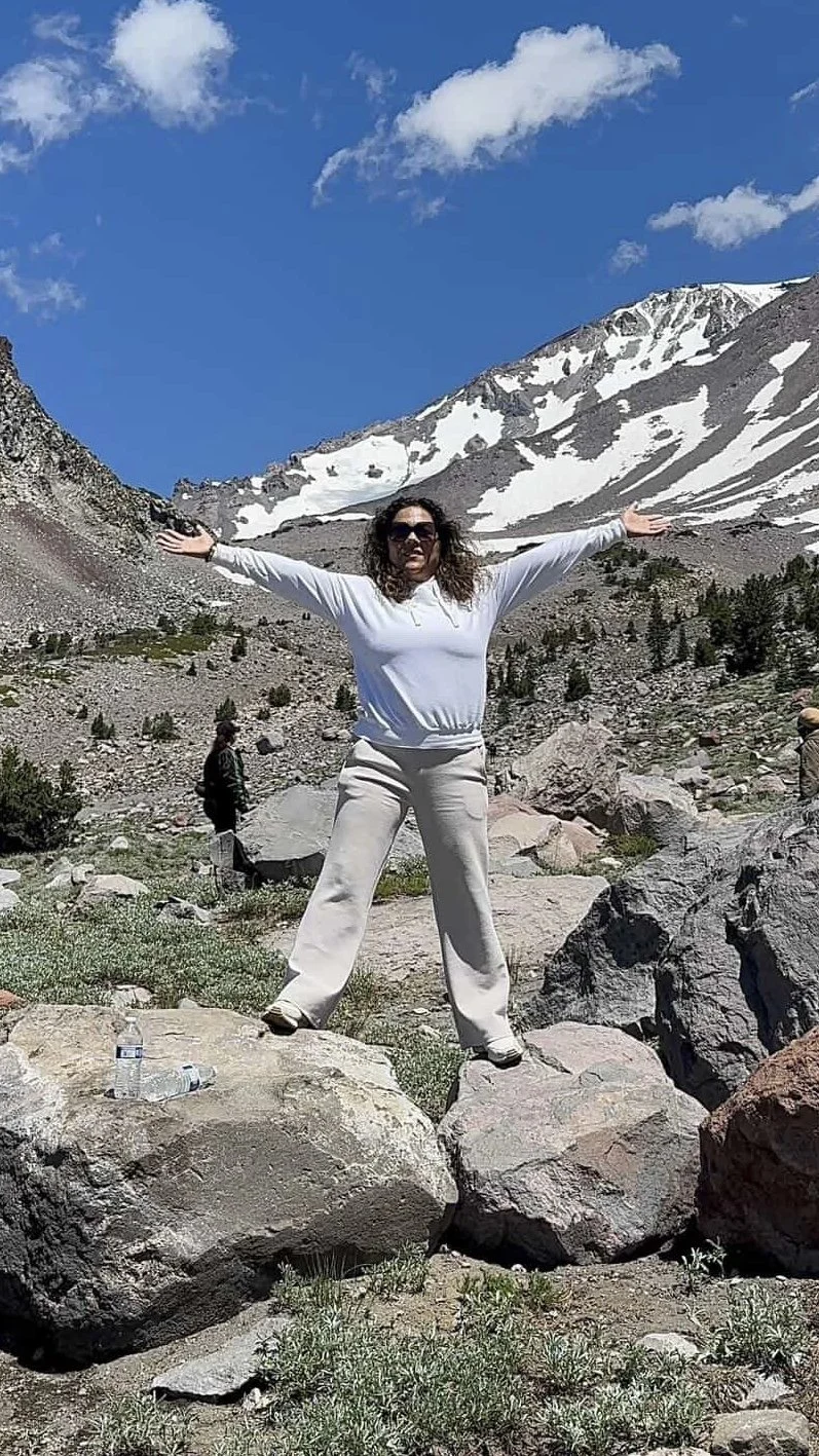 A woman standing on rocks with arms outstretched in front of snow-capped mountains and a clear blue sky.