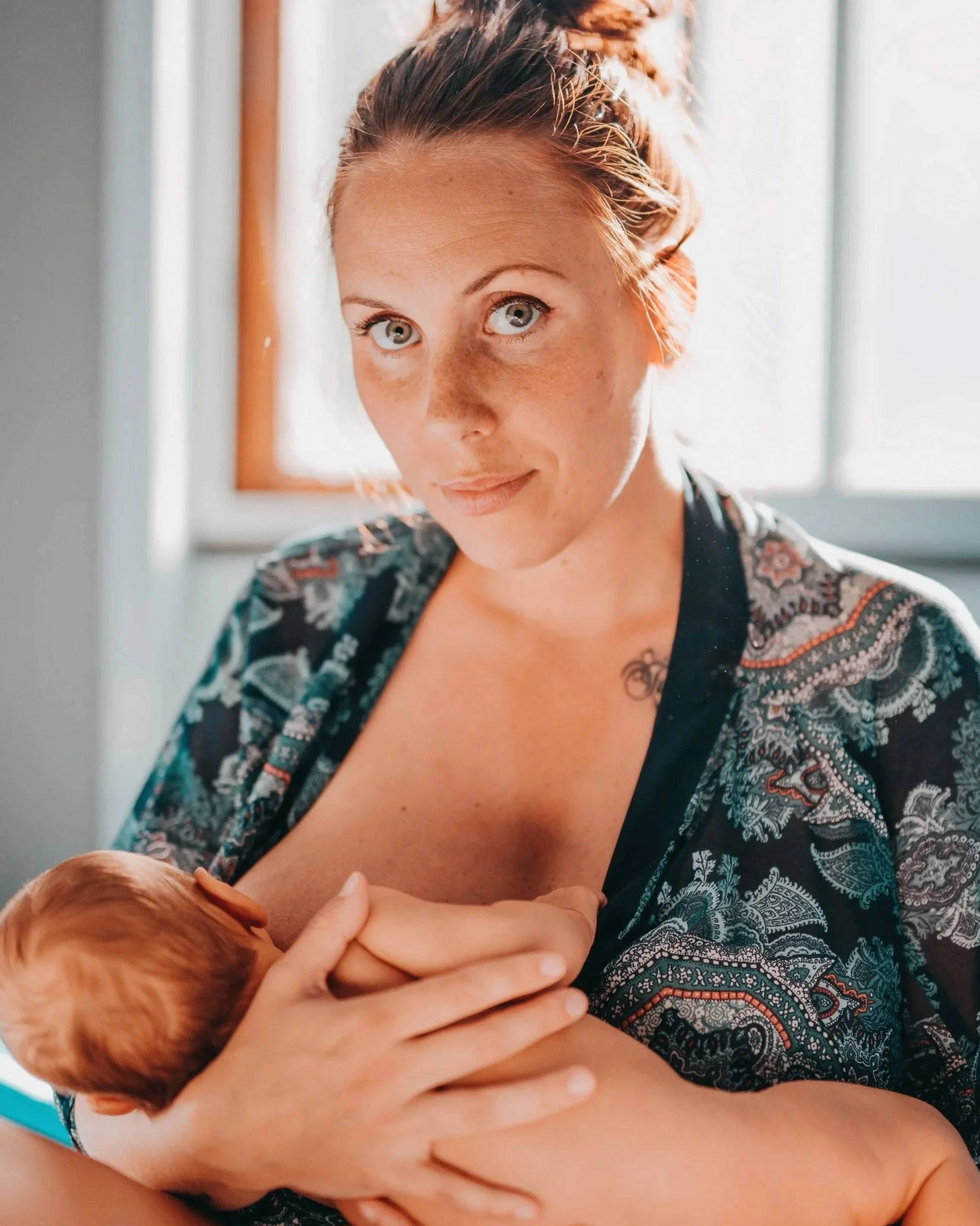 A woman with red hair and freckles holding a newborn with reddish hair close to her chest, with sunlight streaming through the window behind them.
