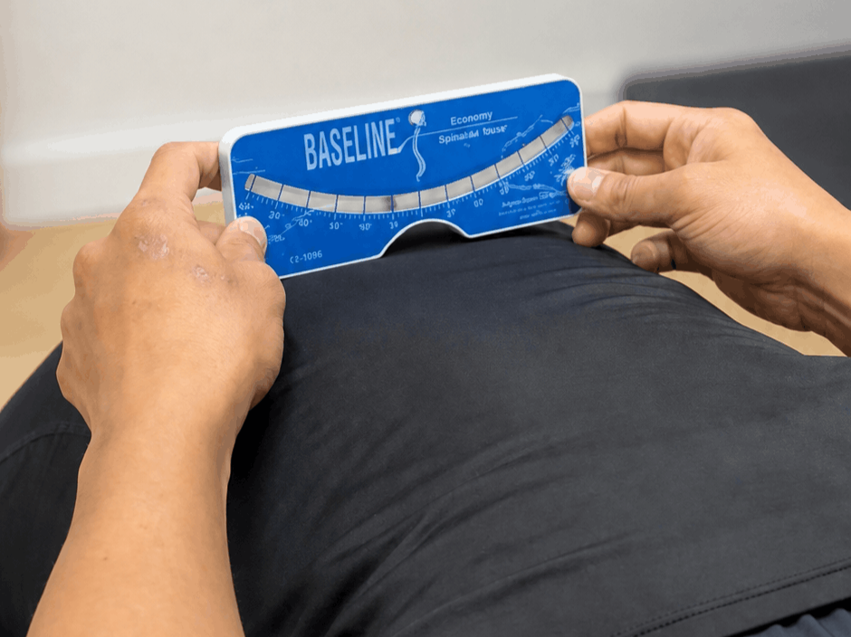 A person lying face down on a medical examination table holds a blue 'Baseline' fitness test card while being in a setting that appears to be a physical therapy or medical office, with medical supplies and furniture in the background.