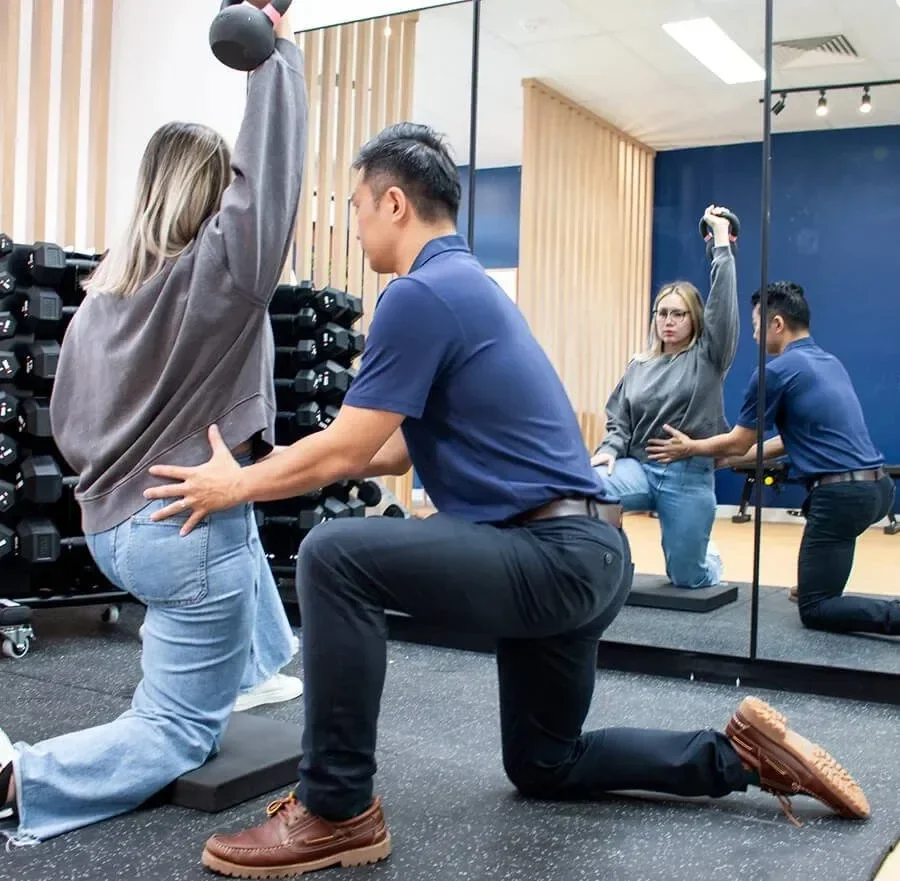 A fitness trainer assisting a woman with a weighted exercise, while another woman practices with a kettlebell in front of a mirror at a gym.