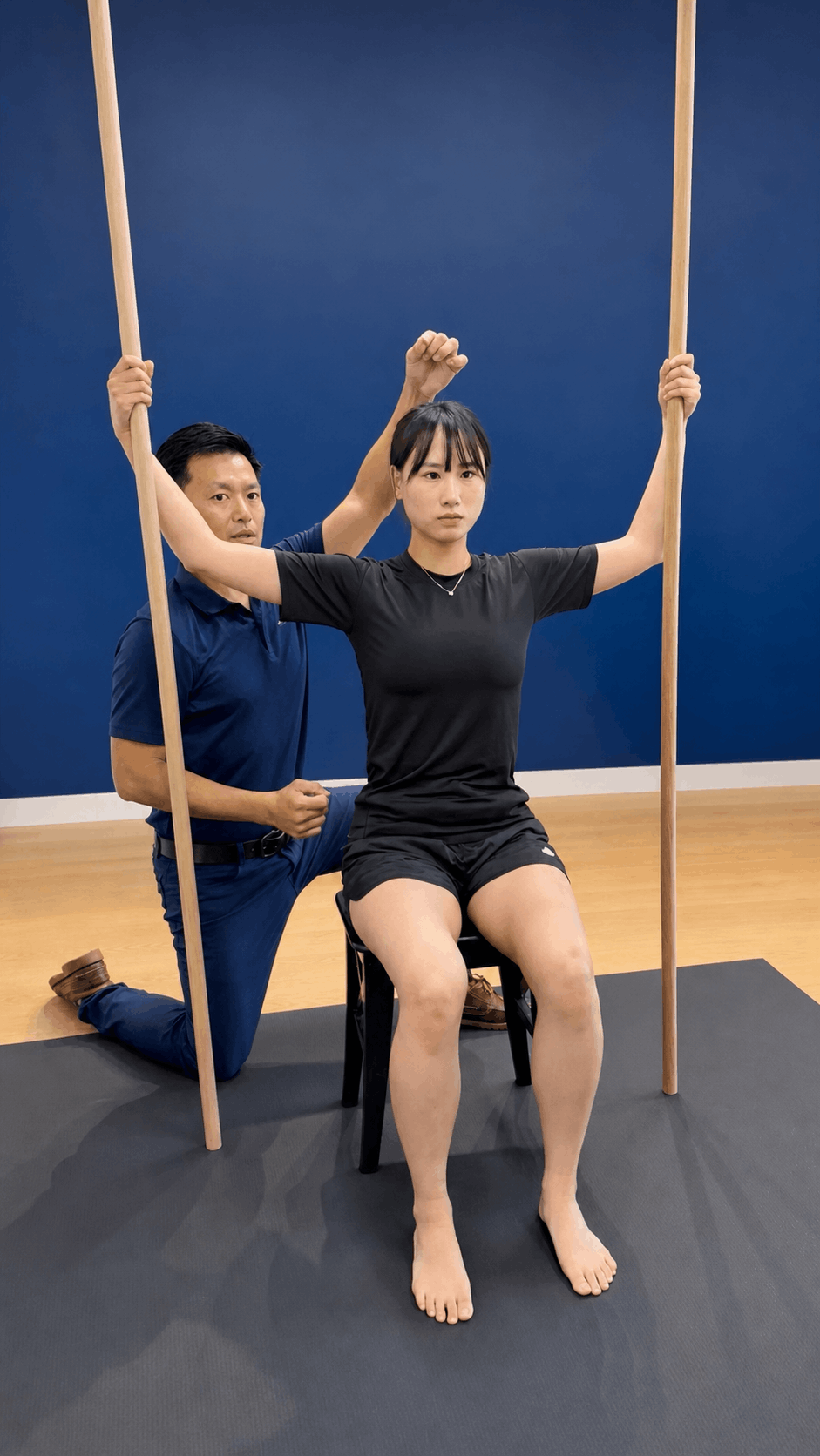 A woman sitting on a chair with her arms extended holding wooden sticks, in a physical therapy or rehabilitation setting. A man kneeling beside her appears to be guiding or assisting her.