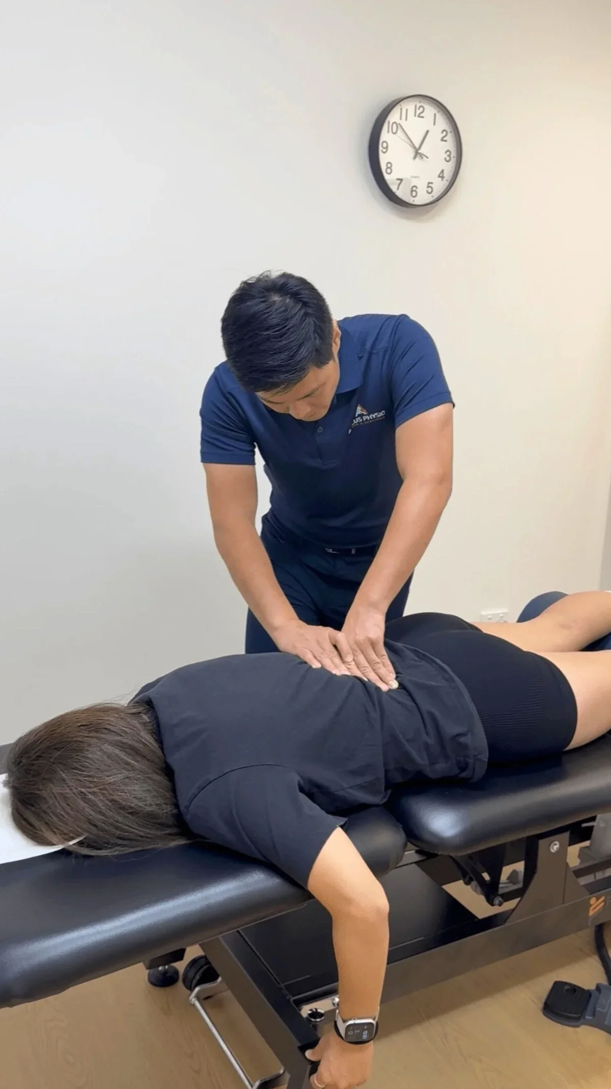 A chiropractor performs an adjustment on a female patient's back in a clinical setting, with a wall clock showing 10:07 in the background.