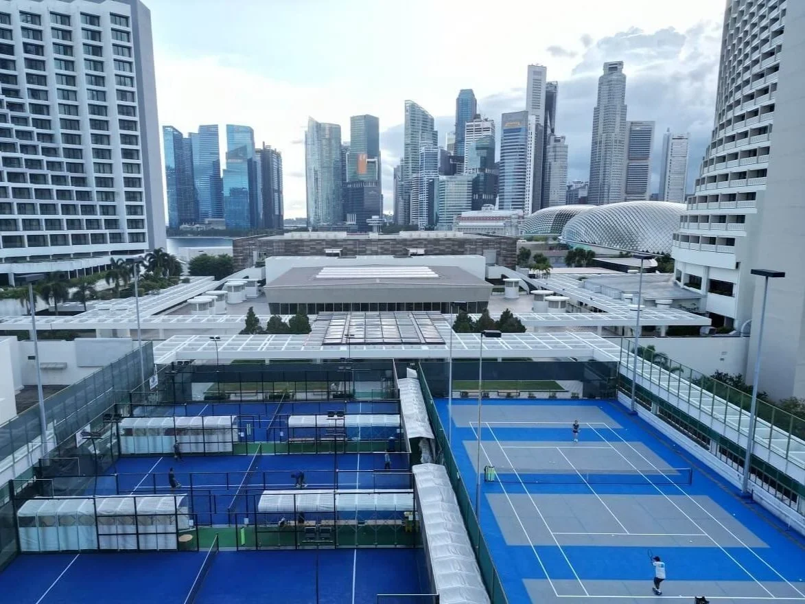 View of outdoor tennis courts with players, surrounded by tall modern skyscrapers in a city skyline.