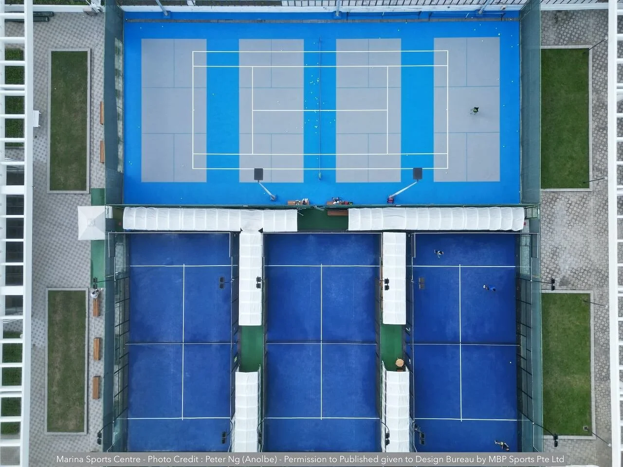 Top-down view of a sports facility with three enclosed tennis courts, two on the bottom and one larger court at the top, with surrounding green areas, benches, and pathways.