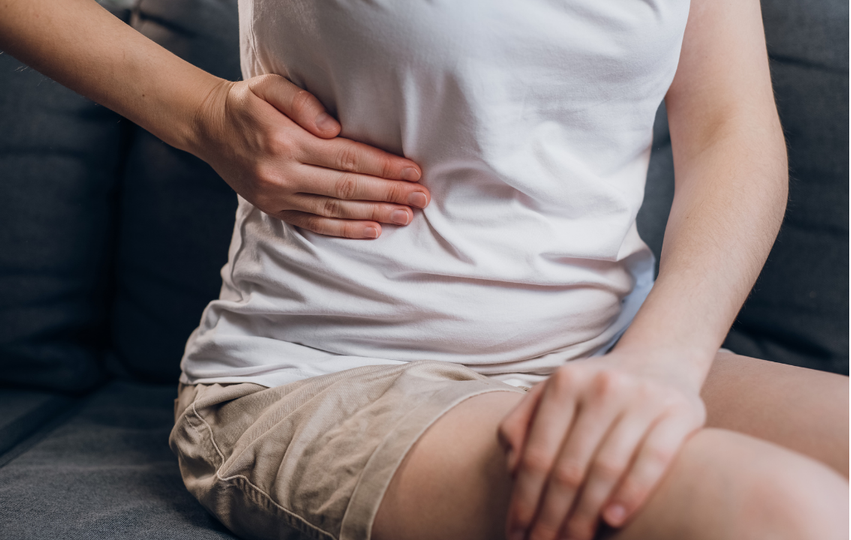 Person sitting on a gray couch holding their stomach with one hand, appearing to experience discomfort or pain.