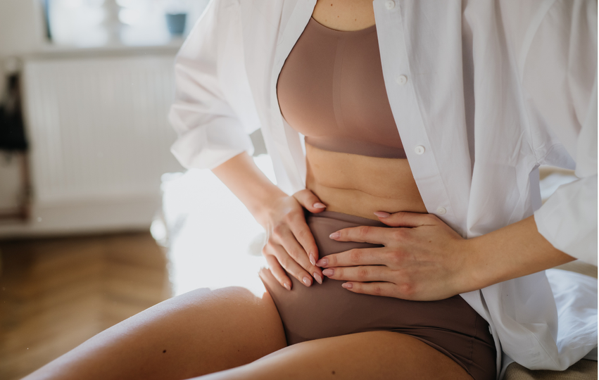 A woman wearing a beige bra and brown underwear, with a white shirt partially open, sitting on a bed with her hands on her stomach in a sunlit room.