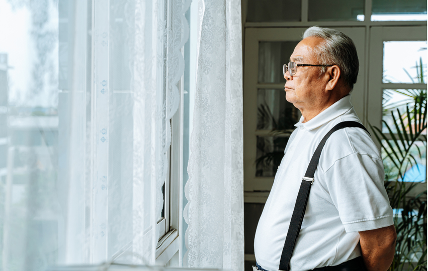 An elderly man with glasses, wearing a white polo shirt and suspenders, standing indoors while looking outside through a window with lace curtains.
