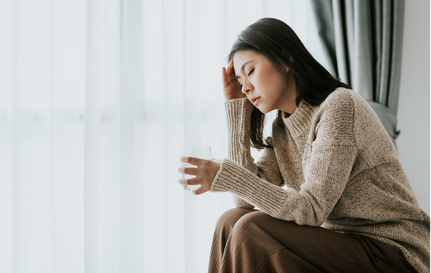 A woman sitting indoors near a window, holding a glass of water, with a look of discomfort or pain on her face.