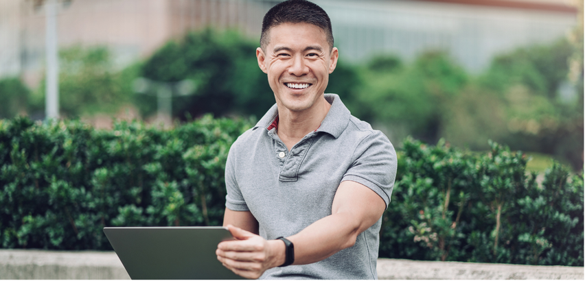 Smiling man in a gray polo shirt sitting outdoors with a laptop in hand, greenery in the background.