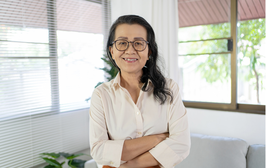 A smiling woman with glasses, wearing a cream-colored blouse, standing with arms crossed in a bright room with large windows and trees outside.