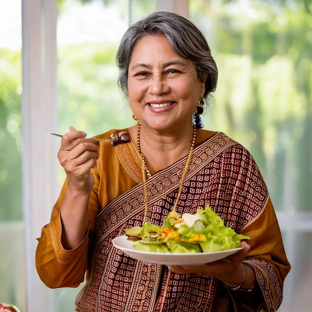 A smiling middle-aged woman with gray hair, wearing traditional Indian attire and jewelry, holding a plate of salad and a fork, standing indoors with a bright, green outdoor background.
