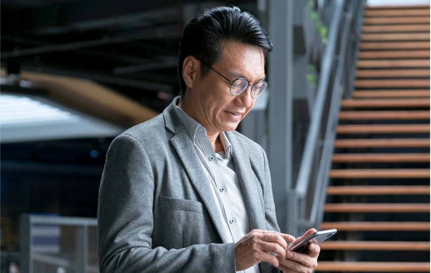 A man with glasses wearing a gray blazer and striped shirt looking at his phone indoors near wooden stairs.