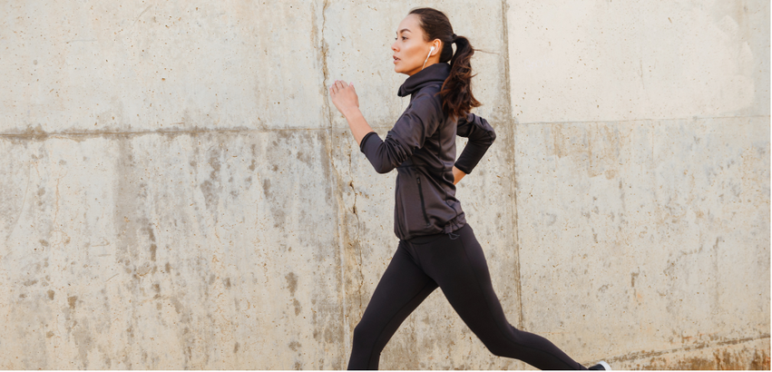 Woman running outdoors near a concrete wall, wearing black athletic clothing and wireless earbuds.
