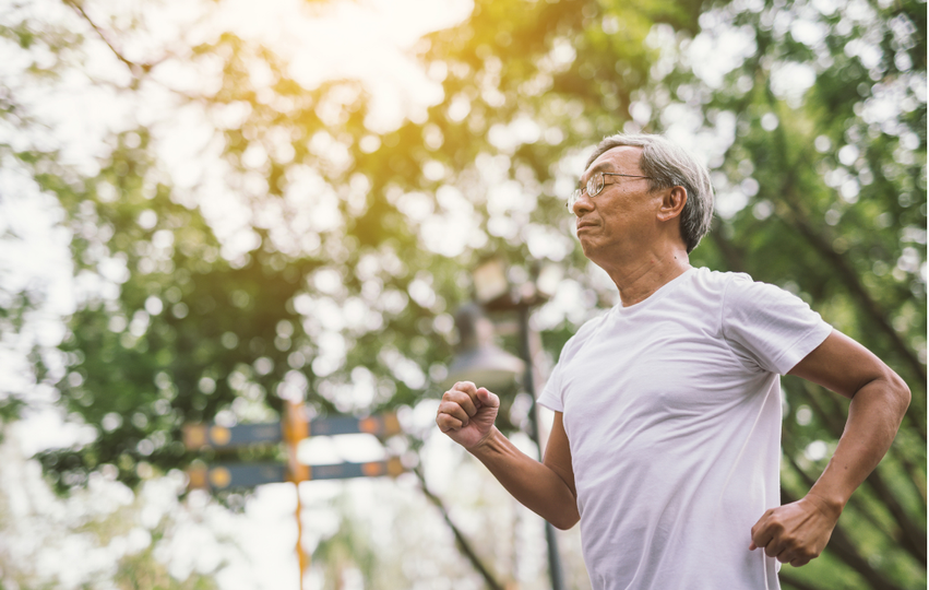 An elderly man running outdoors in a park with trees and sunlight in the background.