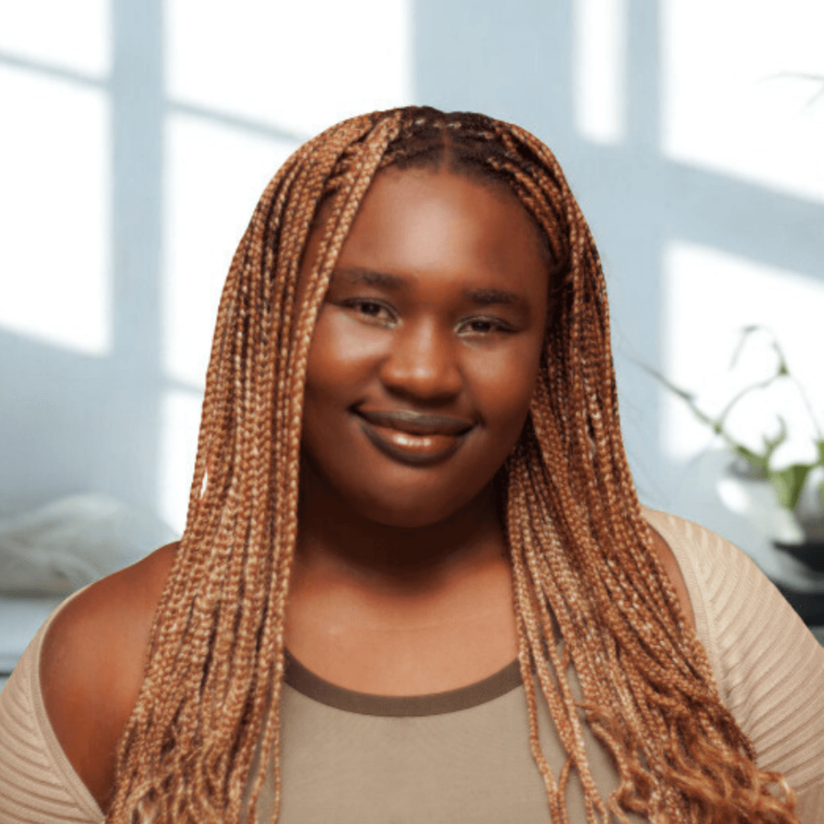 A woman with long, braided hair smiling indoors with sunlight streaming in through the windows.