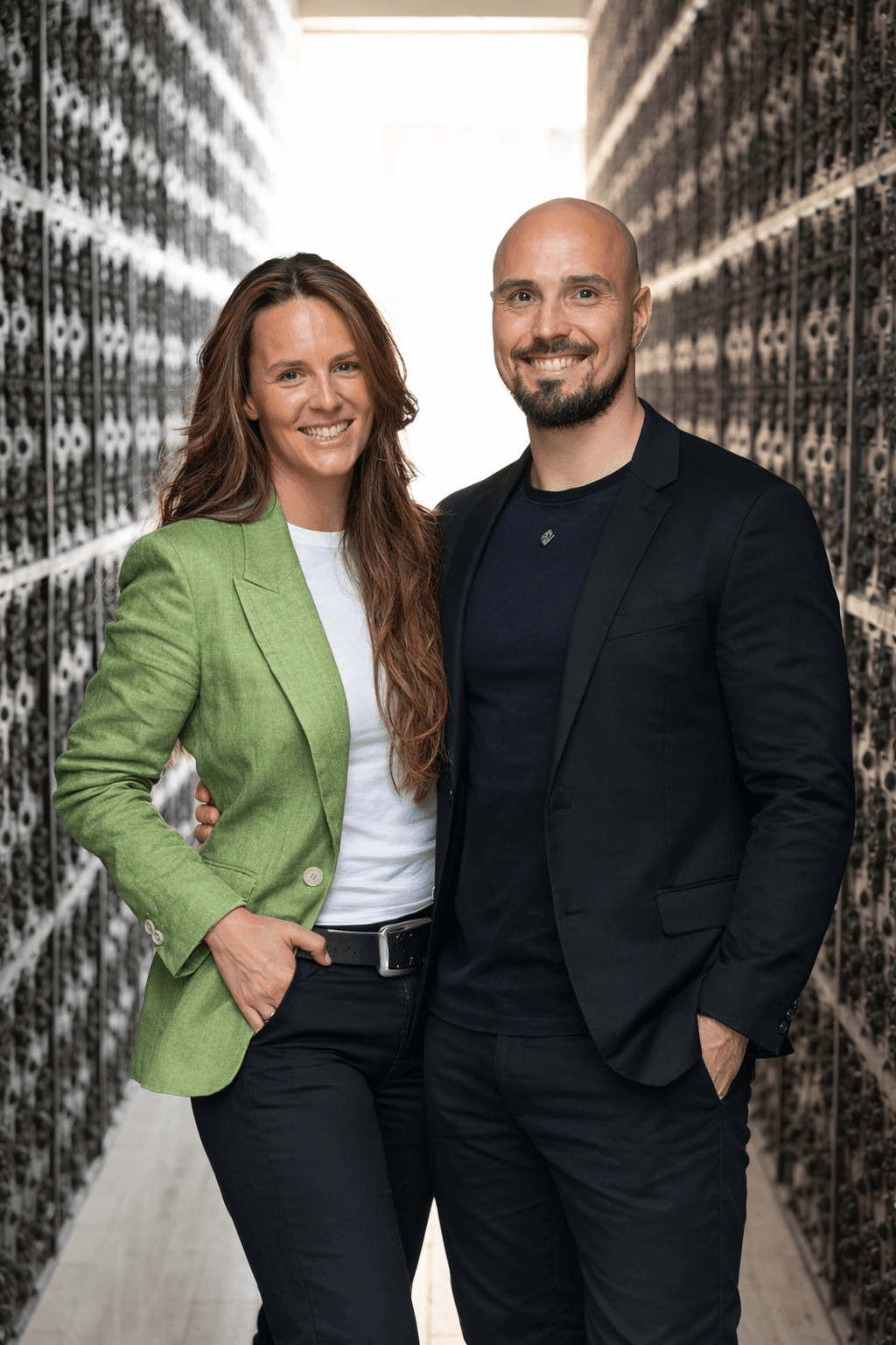 A smiling man and woman standing close together in a narrow aisle with server racks on both sides, in a data center.