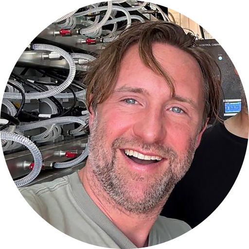 A smiling man with a beard and blue eyes, in front of a server room with numerous cables and equipment.
