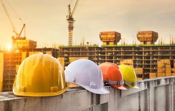 Four construction helmets in yellow, white, orange, and yellow-green are placed on a concrete ledge at a construction site during sunset, with cranes and building frameworks in the background.
