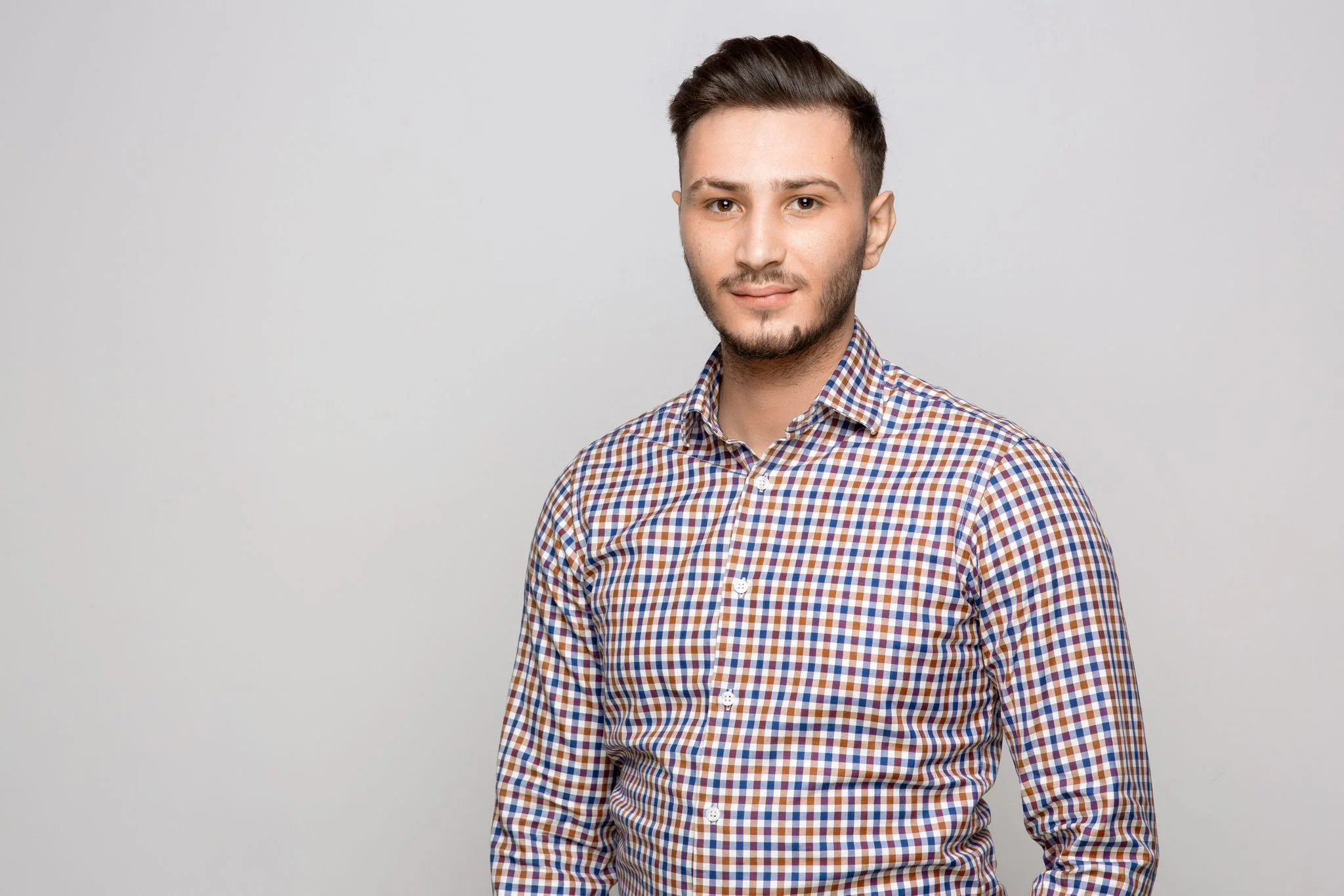 A young man with short dark hair and a beard wearing a checkered shirt standing against a plain white background.