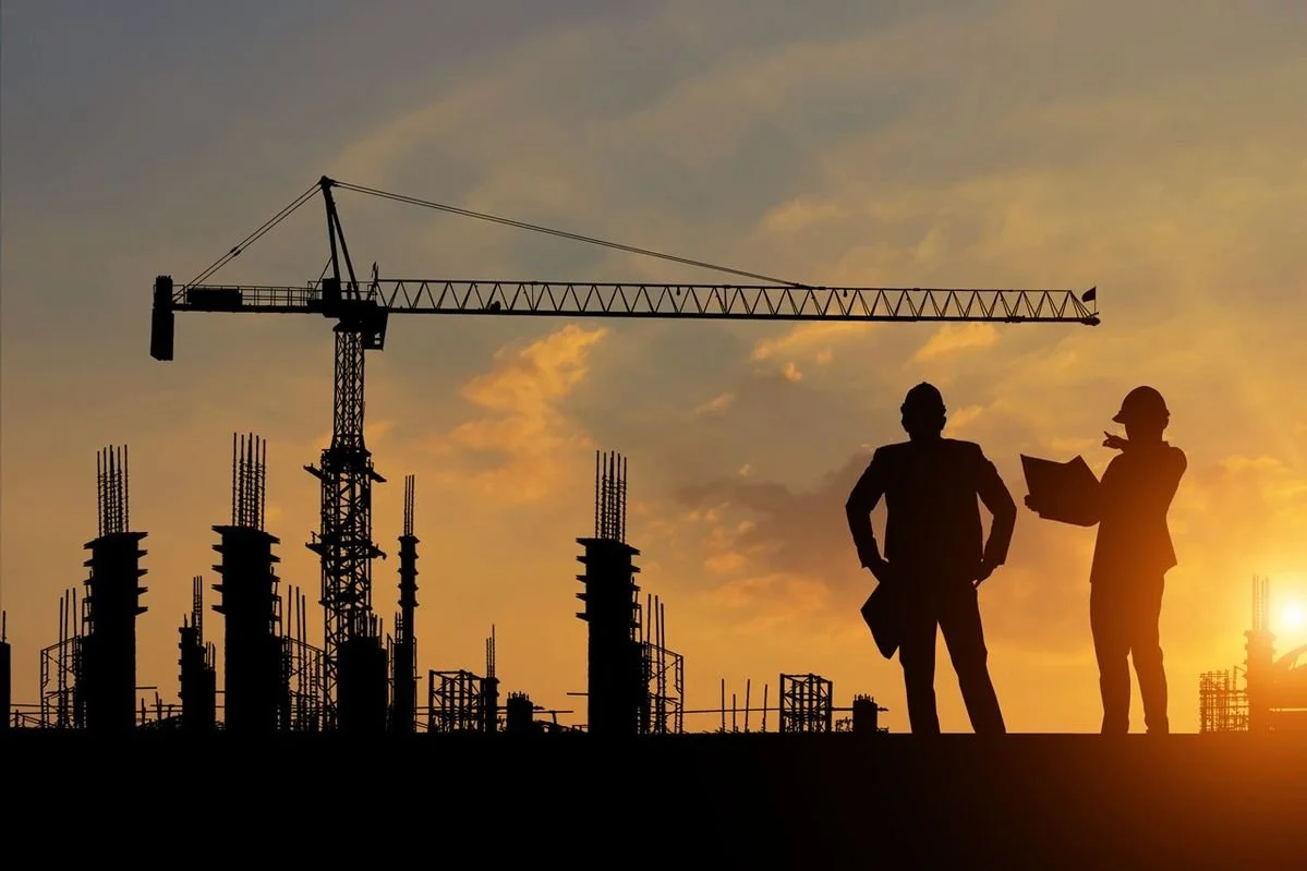 Silhouettes of two construction workers standing on a construction site with steel rebar and scaffolding, a tower crane overhead, during sunset with a partly cloudy sky.