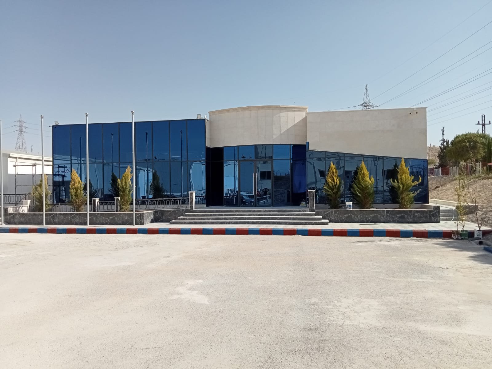 Modern building with blue tinted glass windows, a beige concrete section on top, and small trees in front. Clear sky in the background, with power lines and a sloped hill to the right.