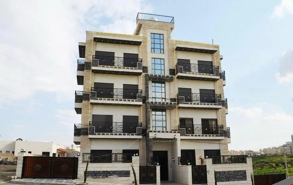 Modern multi-story apartment building with balconies and black railings, beige brick facade, and a rooftop terrace, located in an urban area under a cloudy sky.