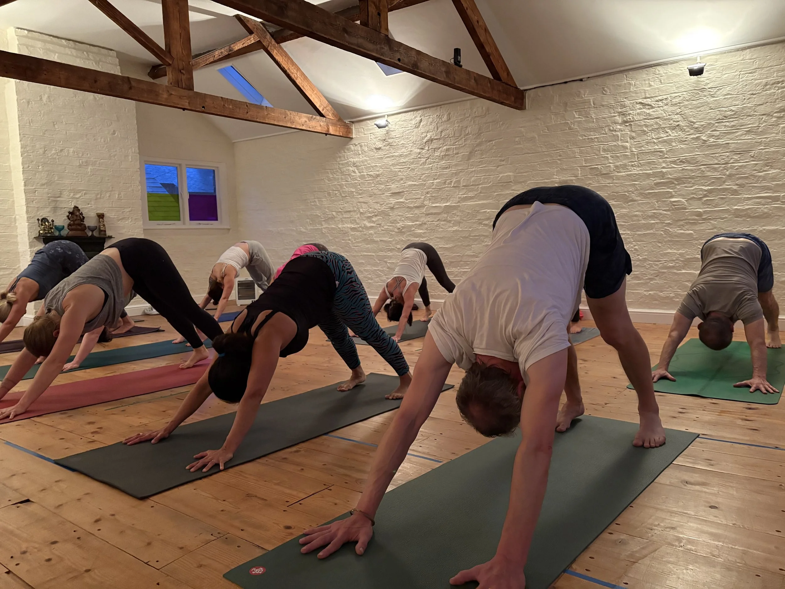 A woman practicing yoga in a push-up position on a black yoga mat in a bright room with wooden floors and white brick walls, with other yoga mats and chairs in the background.