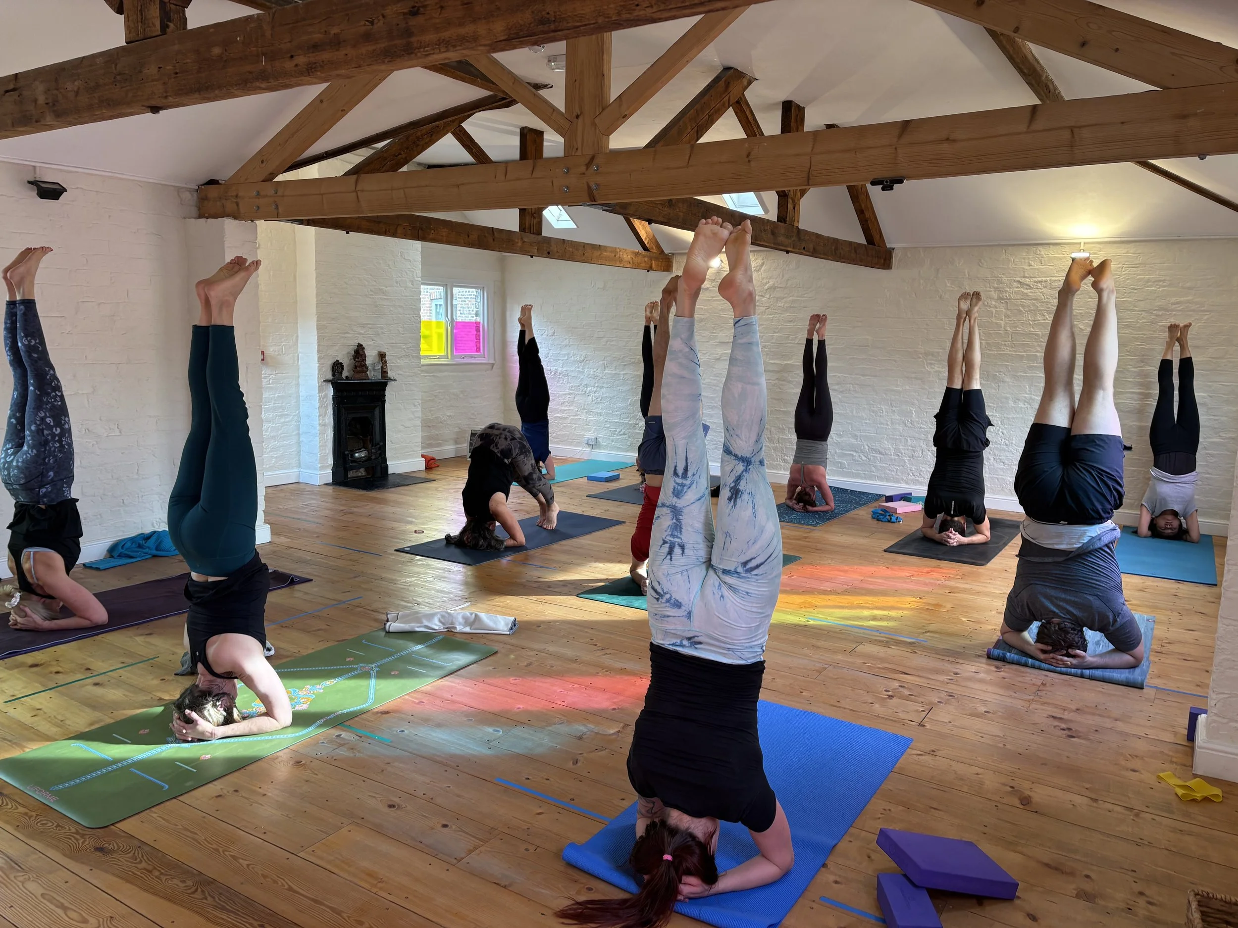 A group of people practicing yoga in a brightly lit yoga studio with white brick walls and wooden floors. They are lying on colorful mats, stretching with one leg raised and held with a strap, while an instructor guides them.