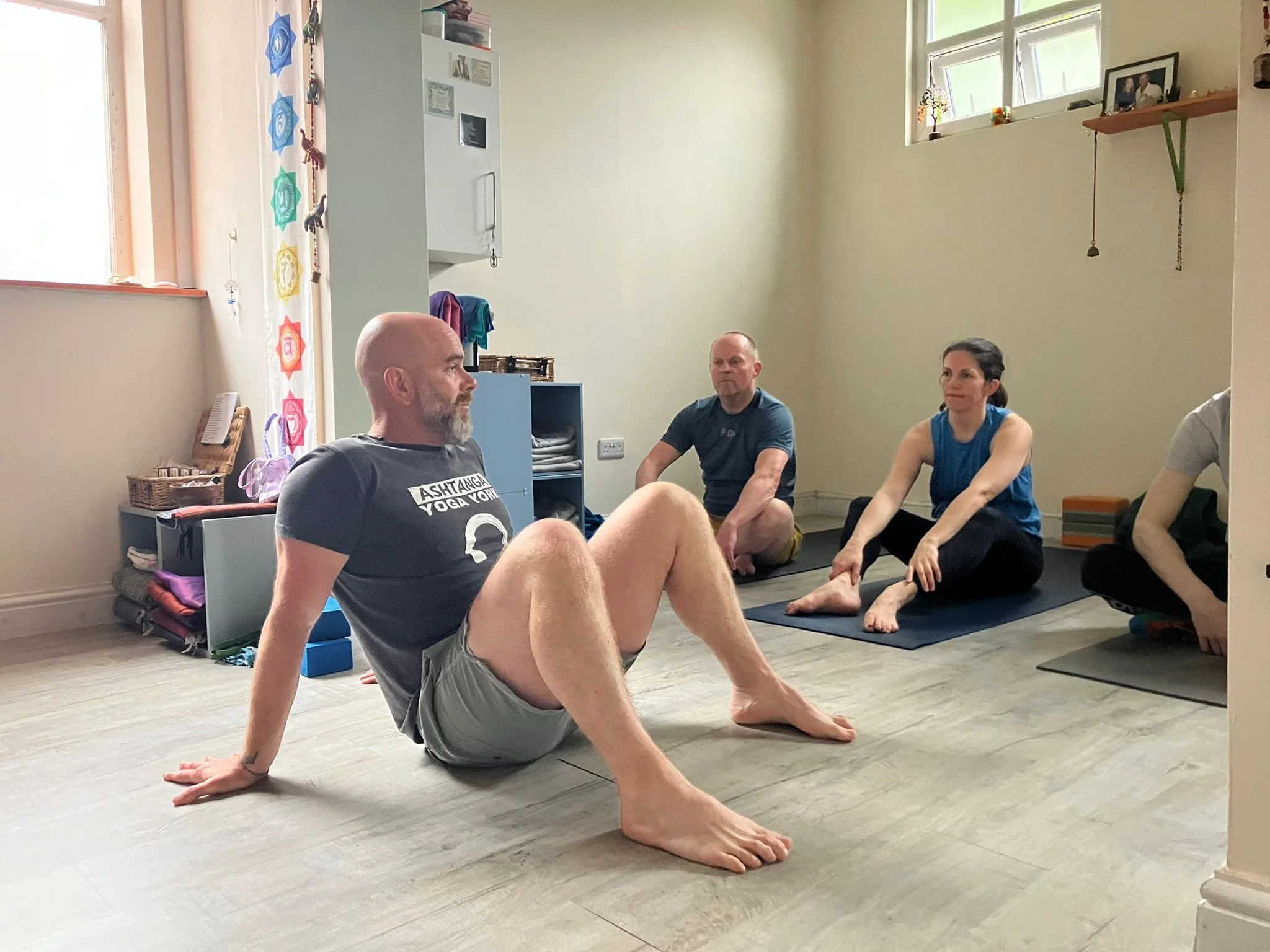 A woman practicing yoga on a mat with a support block, stretching towards a man sitting cross-legged, who is holding her hands. Other participants are in the background doing yoga.