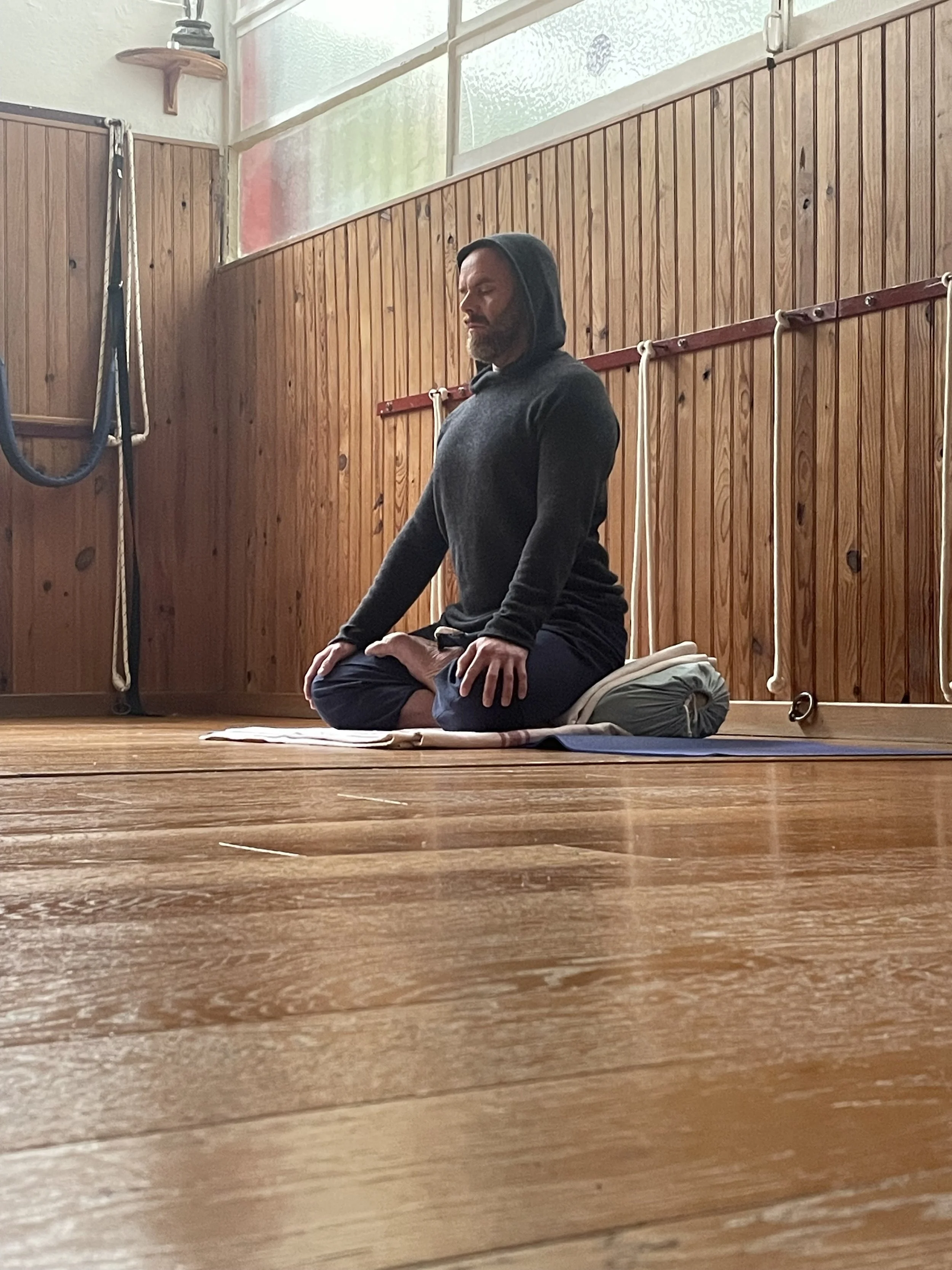 A man sitting in lotus position doing breath work. The room has wooden floors and Iyengar ropes on the walls.