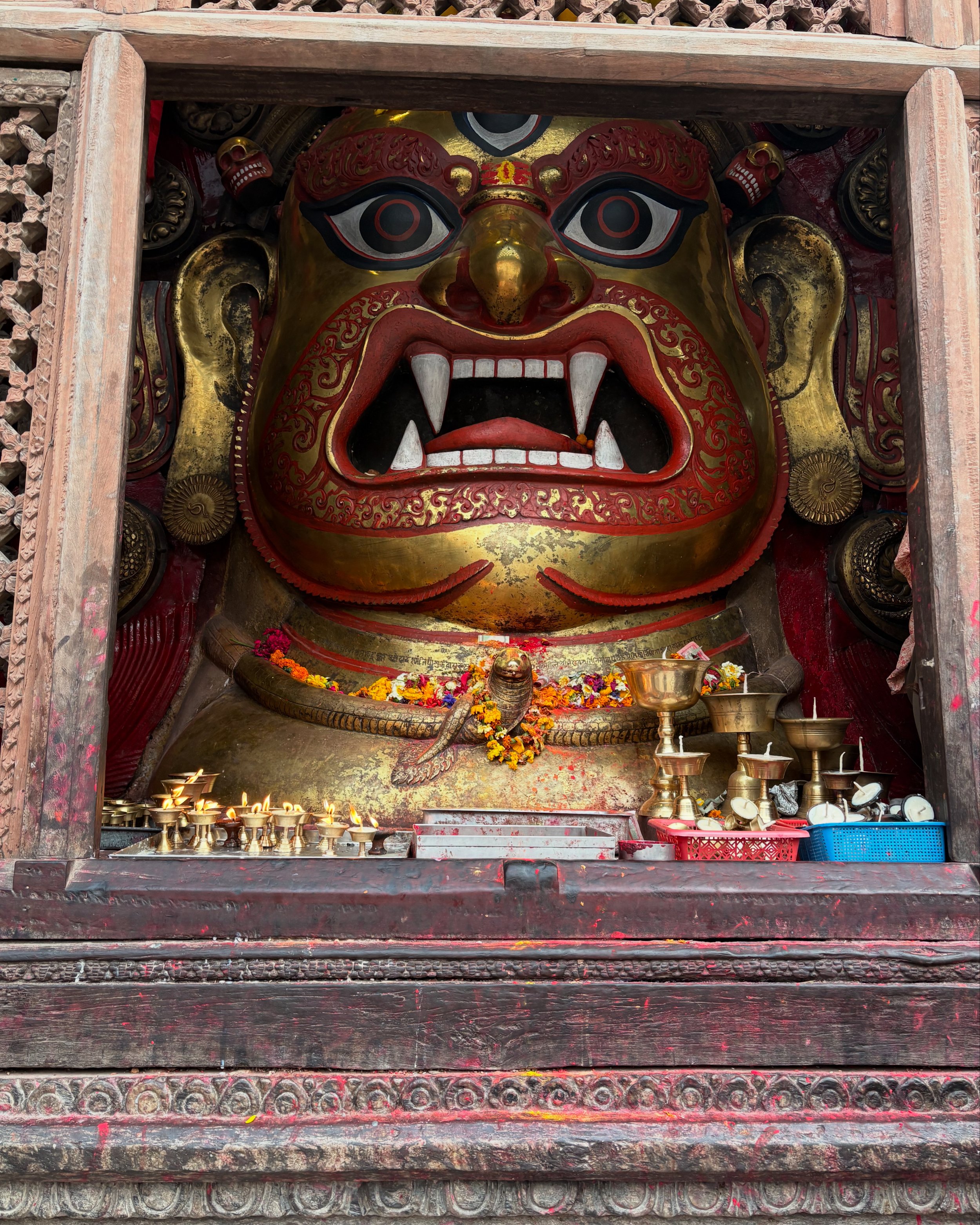 A large decorative mask of a fierce face with bulging eyes, red and gold colors, and sharp teeth, likely from Nepal or Tibet. The mask is housed within a wooden frame and surrounded by offerings, flowers, and candles.