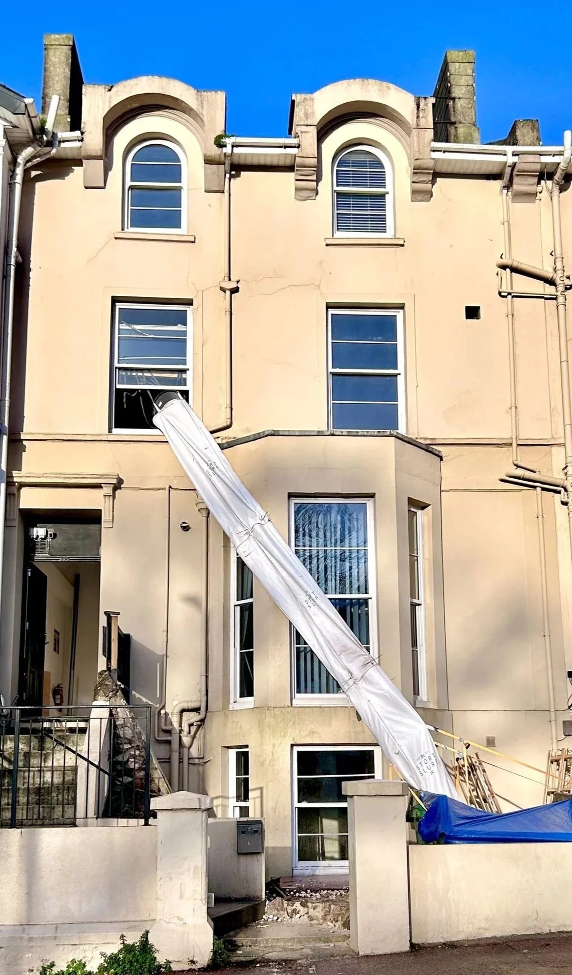 A multi-story beige residential building with several windows, a black railing, and a construction tarp covering a section of the stairs, with bright blue sky in the background.