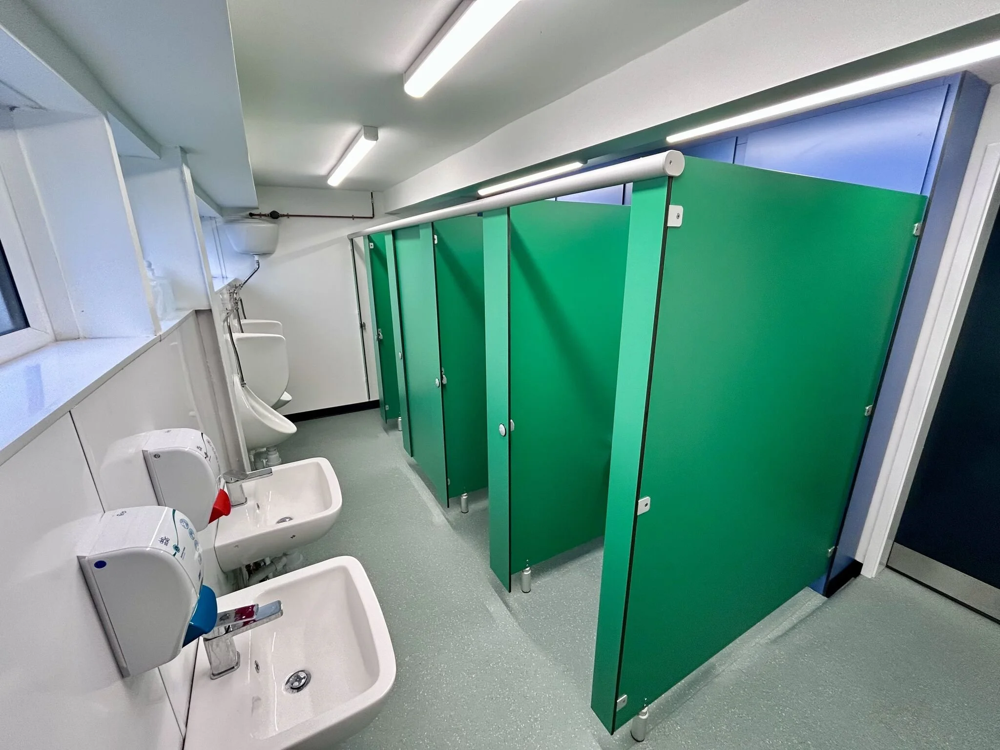 A public restroom with three urinals and green bathroom stalls.