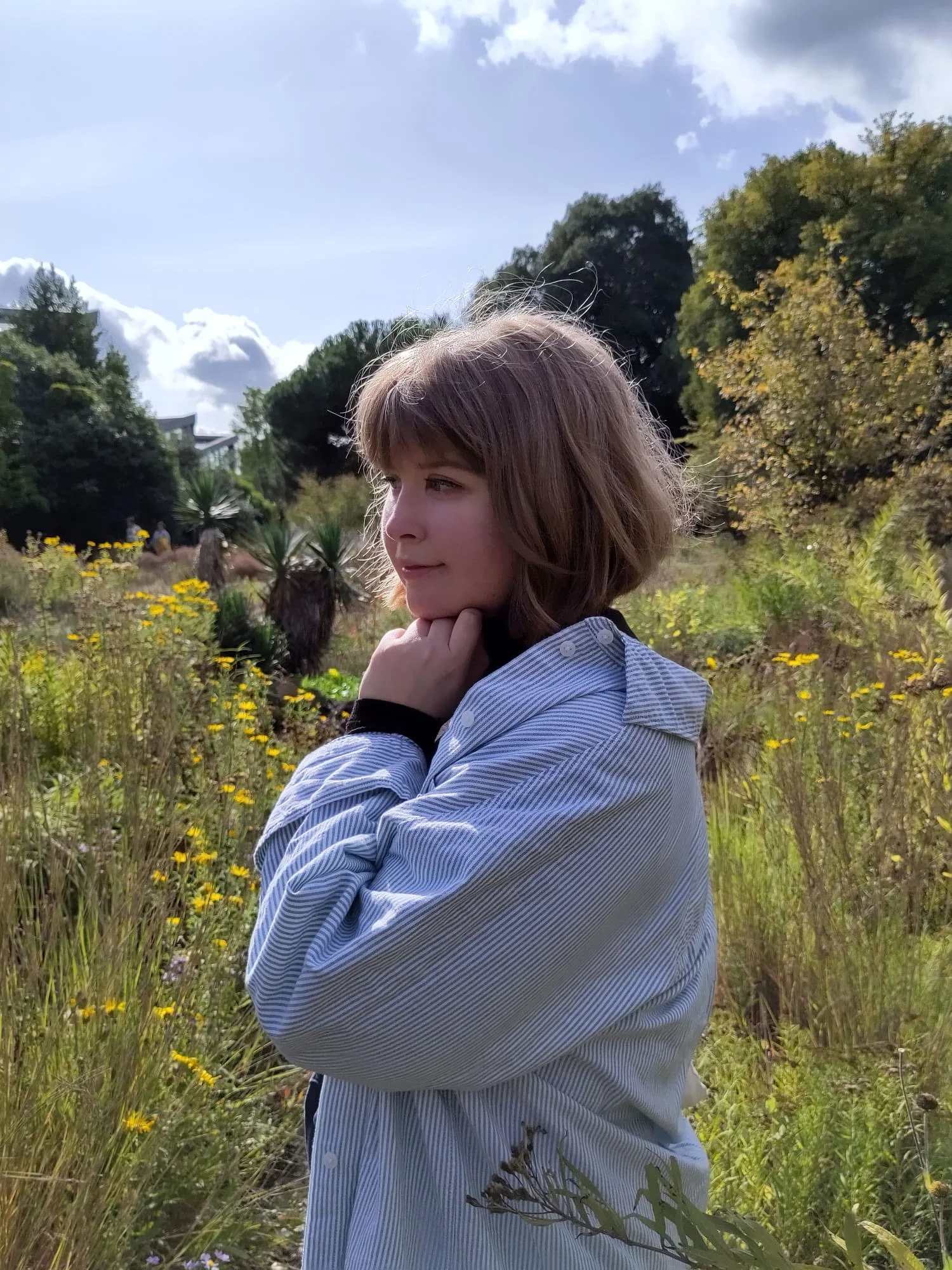 Young woman with a blond bob haircut standing outdoors in a field of wildflowers, looking to the side with a thoughtful expression under a partly cloudy sky.