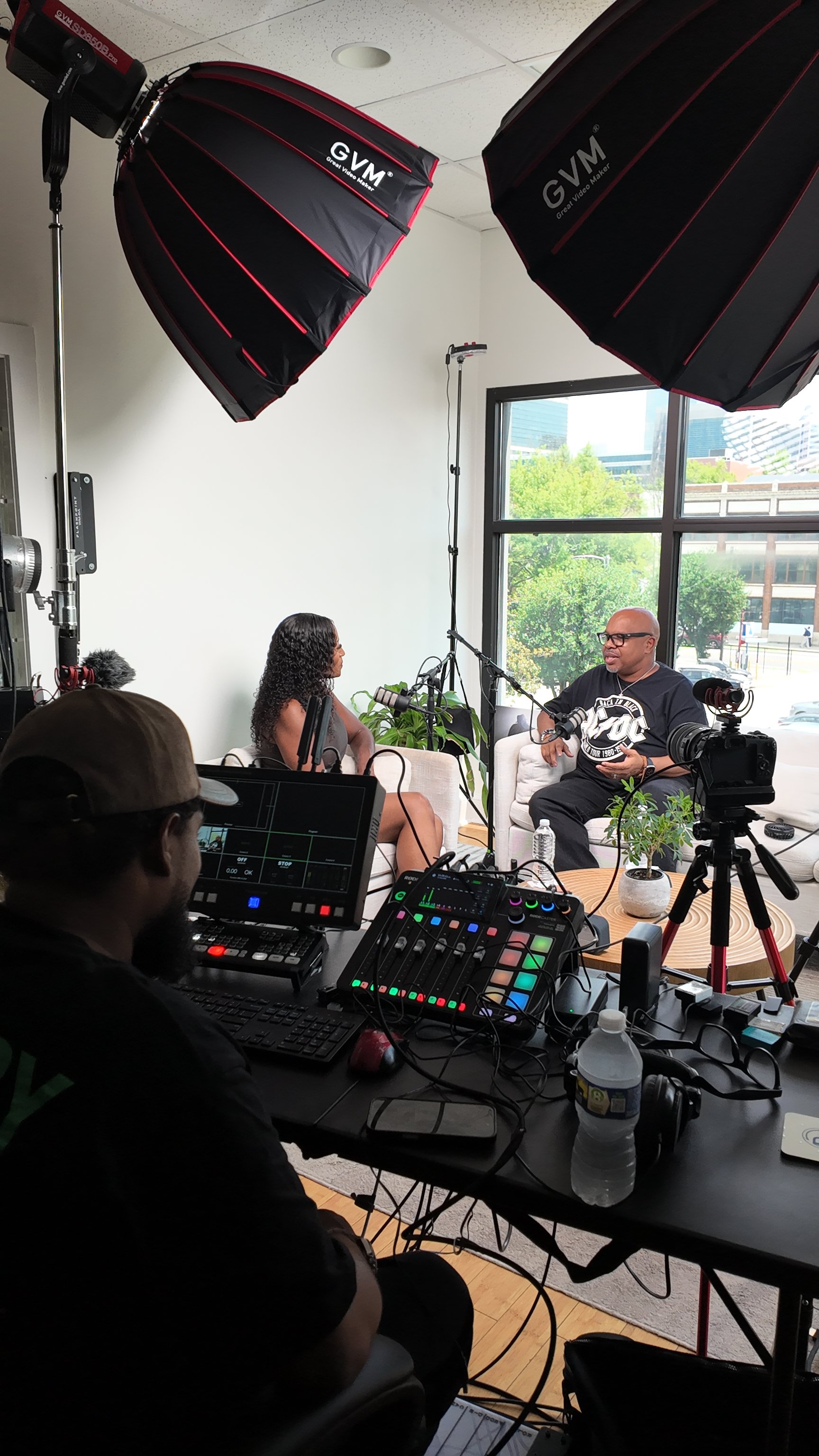Two people sit on white couches having a conversation during an interview or recording session in a well-lit room with large windows. Video and audio equipment, including cameras, microphones, and lighting, are set up around them. A person operating a control panel and monitor is in the foreground, managing the recording.