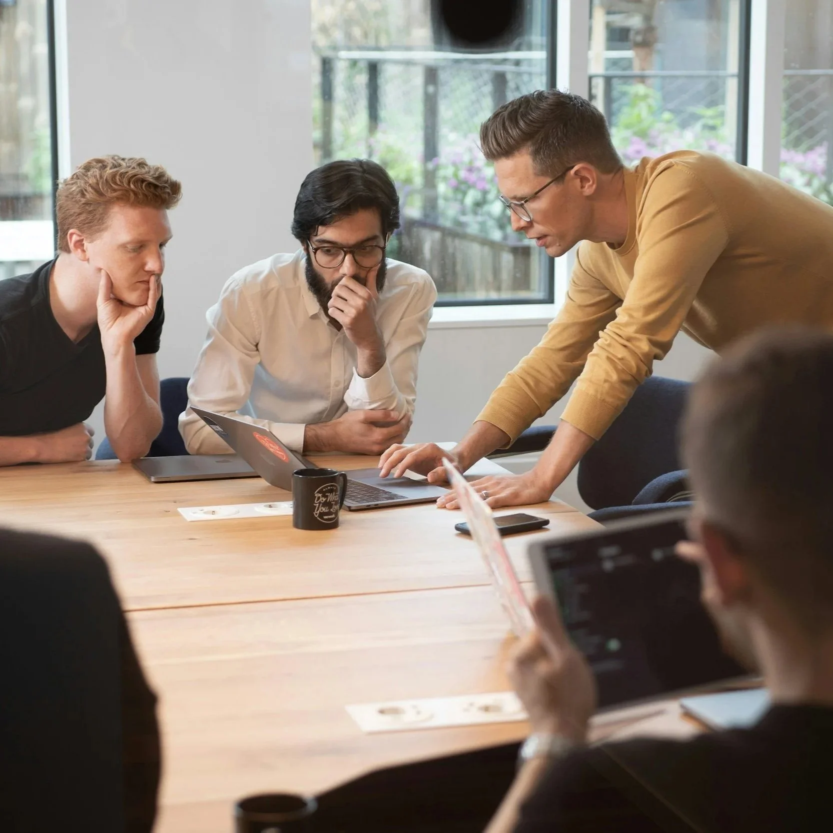 A group of five people in a meeting room, with one person standing and presenting in front of a laptop, while others are sitting and listening attentively, with some looking at their devices.