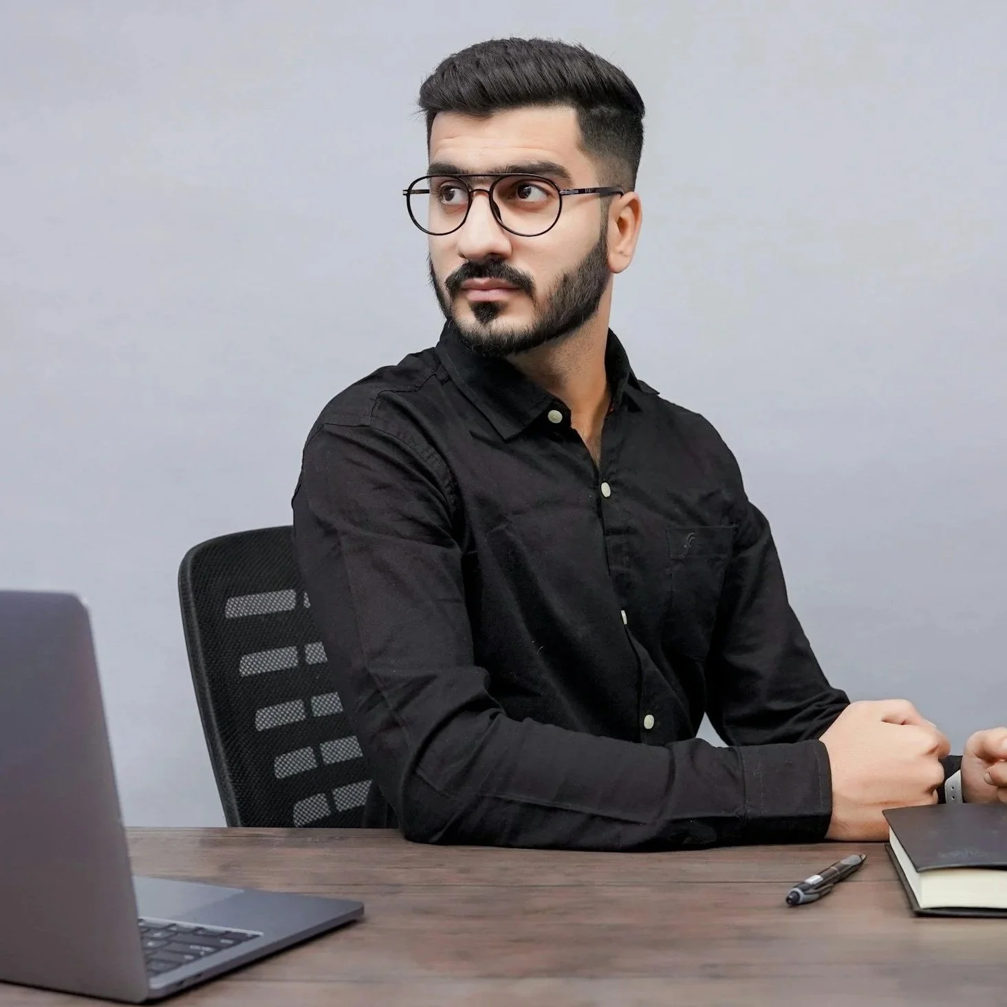 A man with dark hair, beard, glasses, wearing a black shirt, sitting at a desk with a laptop, notebook, and pen, looking to the side against a plain grey wall background.