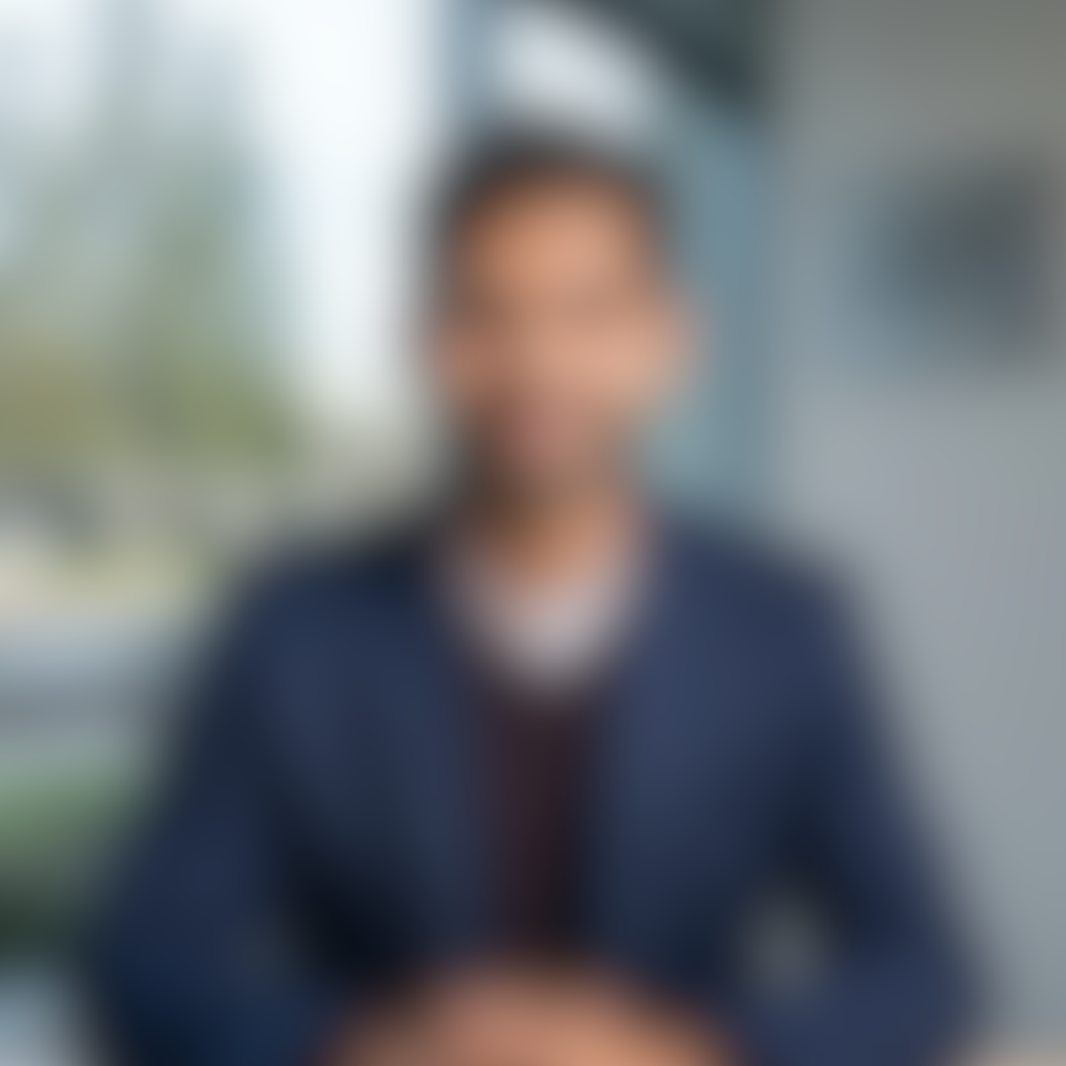 Blurred image of a person sitting indoors near a window, wearing a dark blazer.