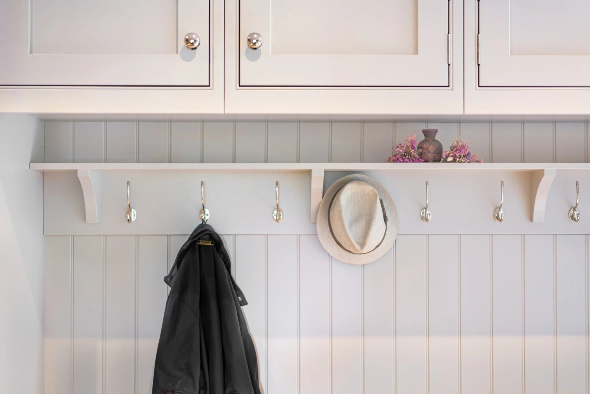 A white coat rack with brass hooks, a straw hat hanging on one hook, and a black jacket hanging on another hook, mounted on a wall with white beadboard paneling. Above the coat rack is a white shelf with a decorative vase and some pink flowers. Part of a white cabinet with round knobs is visible at the top.
