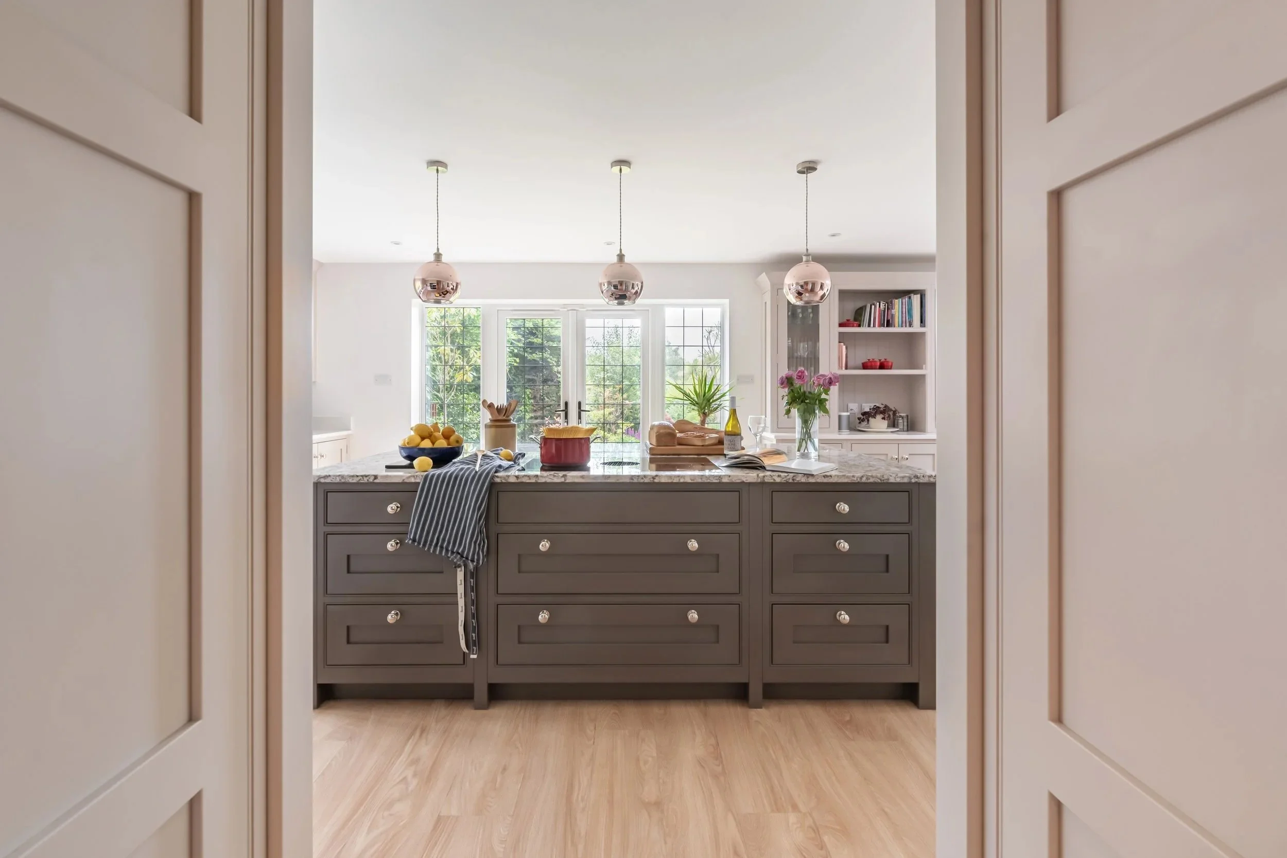 View of a modern kitchen through a doorway, featuring a gray island with various items including lemons, a wine bottle, flowers, and bread. Above, three metallic pendant lights hang from the ceiling. In the background, large windows let in natural light and show a view of greenery outside.