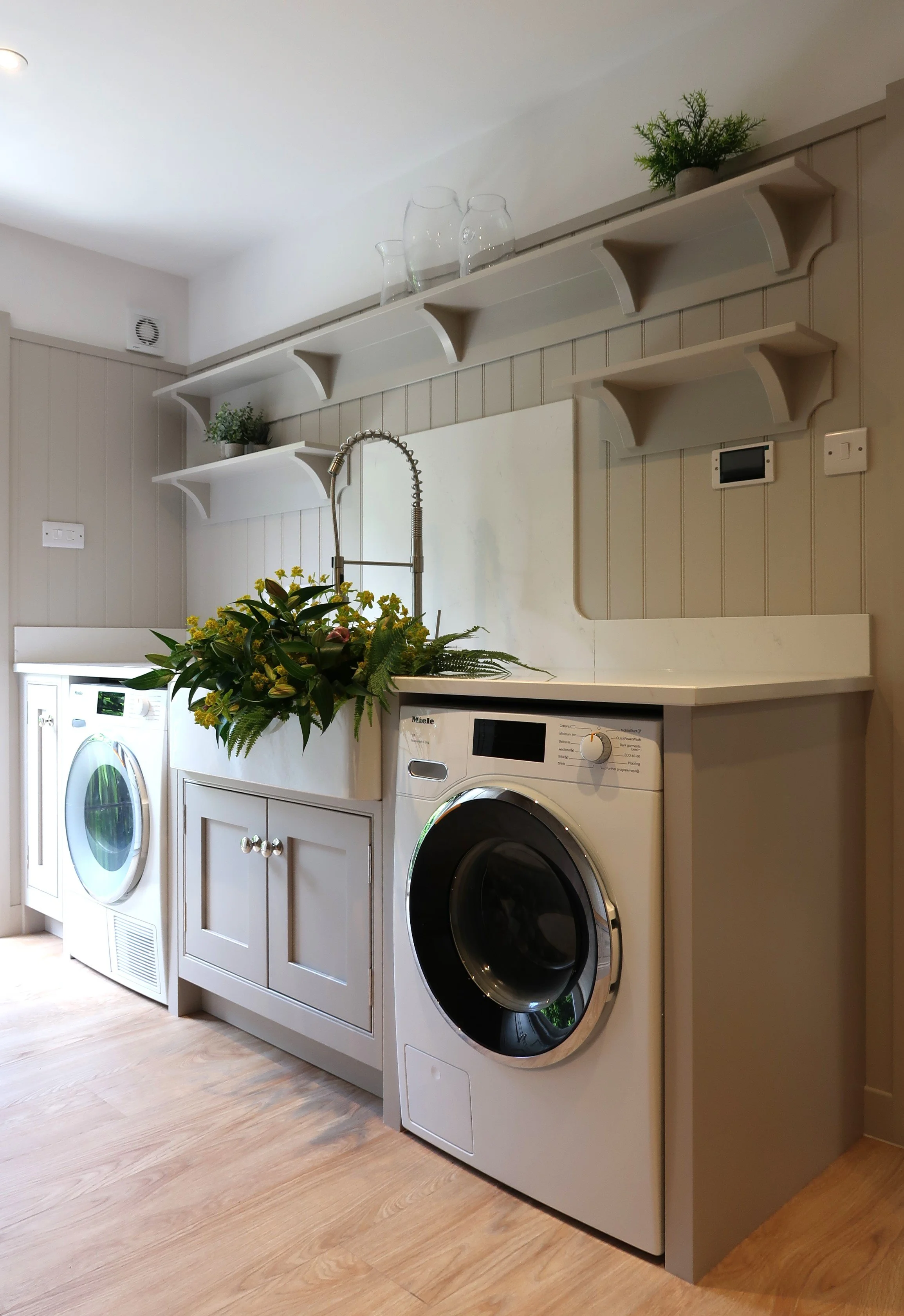 Laundry room with white cabinets, a washing machine, a dryer, and a countertop with a green plant. Shelves above hold glass pitchers and a small potted plant.