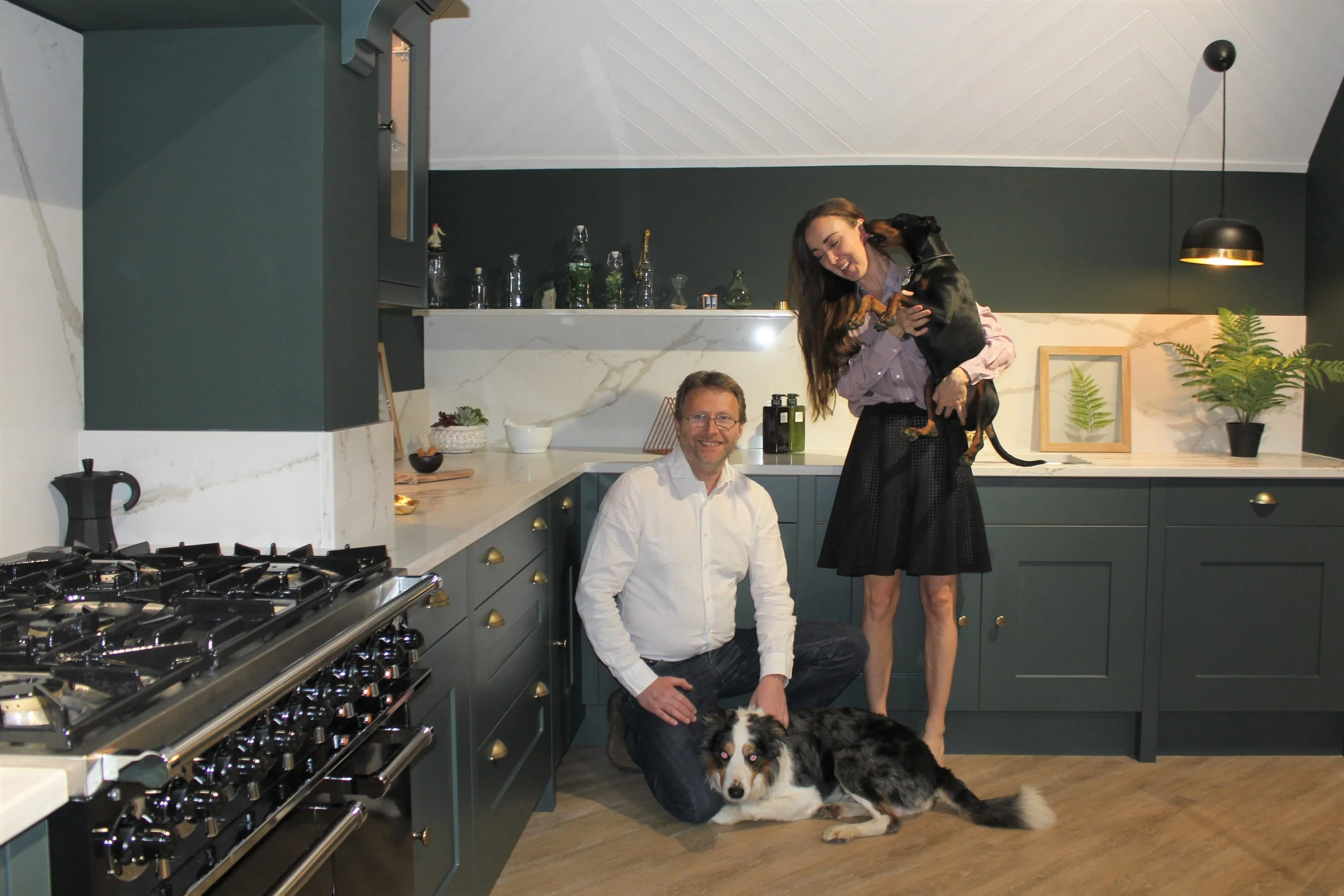 A man kneeling on the kitchen floor with a large dog lying in front of him. A woman is standing next to him, holding a smaller dog in her arms, smiling and engaging with the dogs. The kitchen features dark green cabinets, a marble backsplash, and a countertop with various bottles and plants.