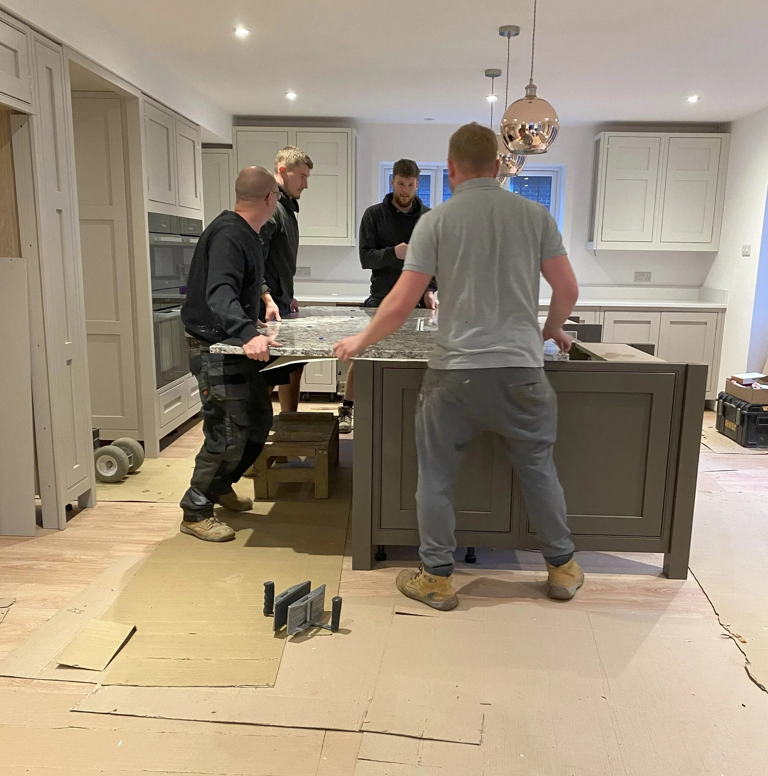 Four men standing around kitchen island in a well-lit modern kitchen, engaged in conversation, with construction tools and materials on the floor and countertop, indicating ongoing kitchen renovation or work.