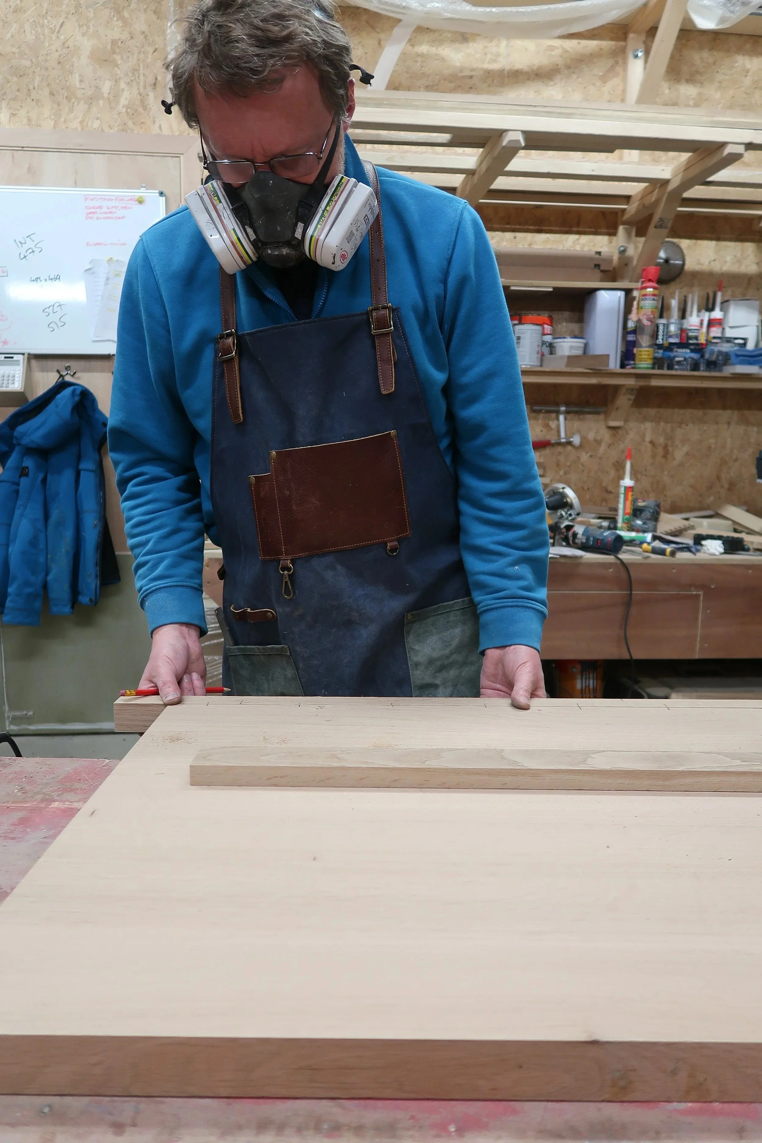 A man in a workshop, wearing safety glasses and a respirator, standing and working on a piece of wood on a workbench.